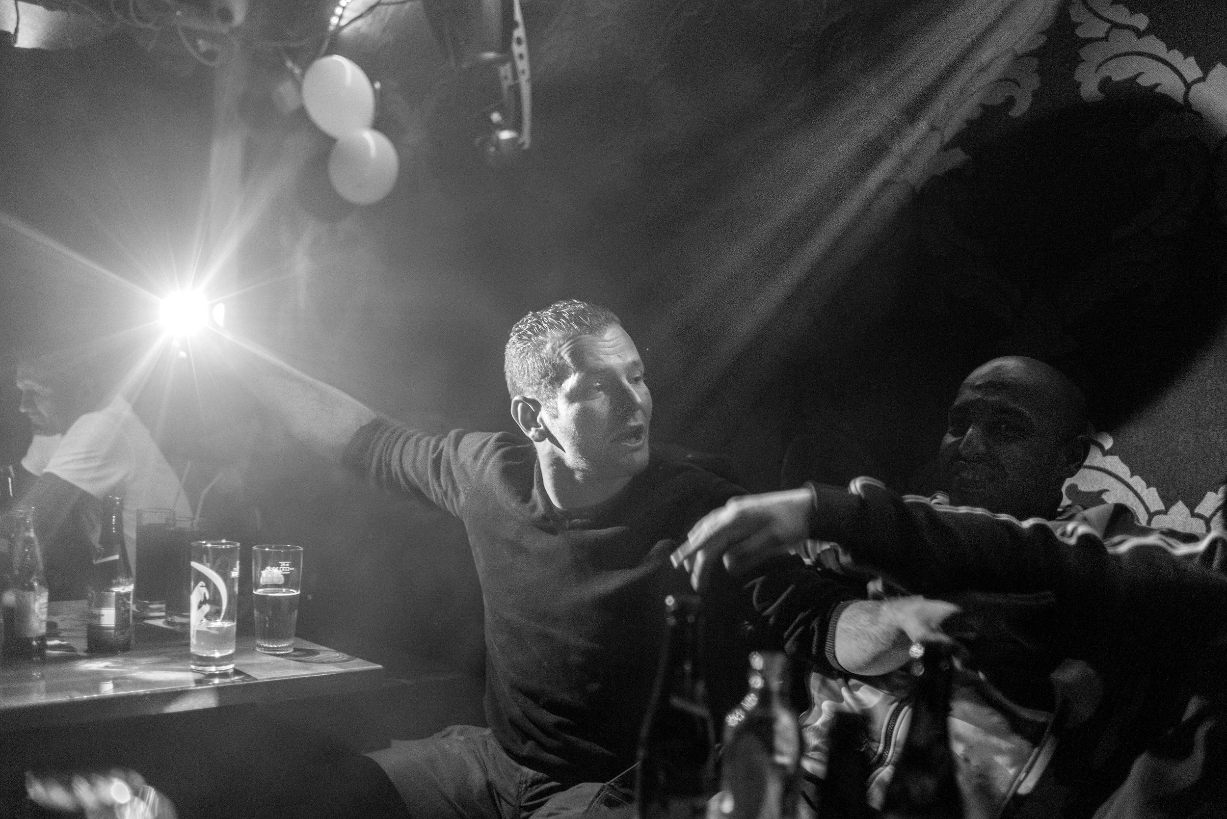 Twee mannen die dansen en elkaar vasthouden in een donkere club met ballonnen en drank op tafel, zwart-wit foto.