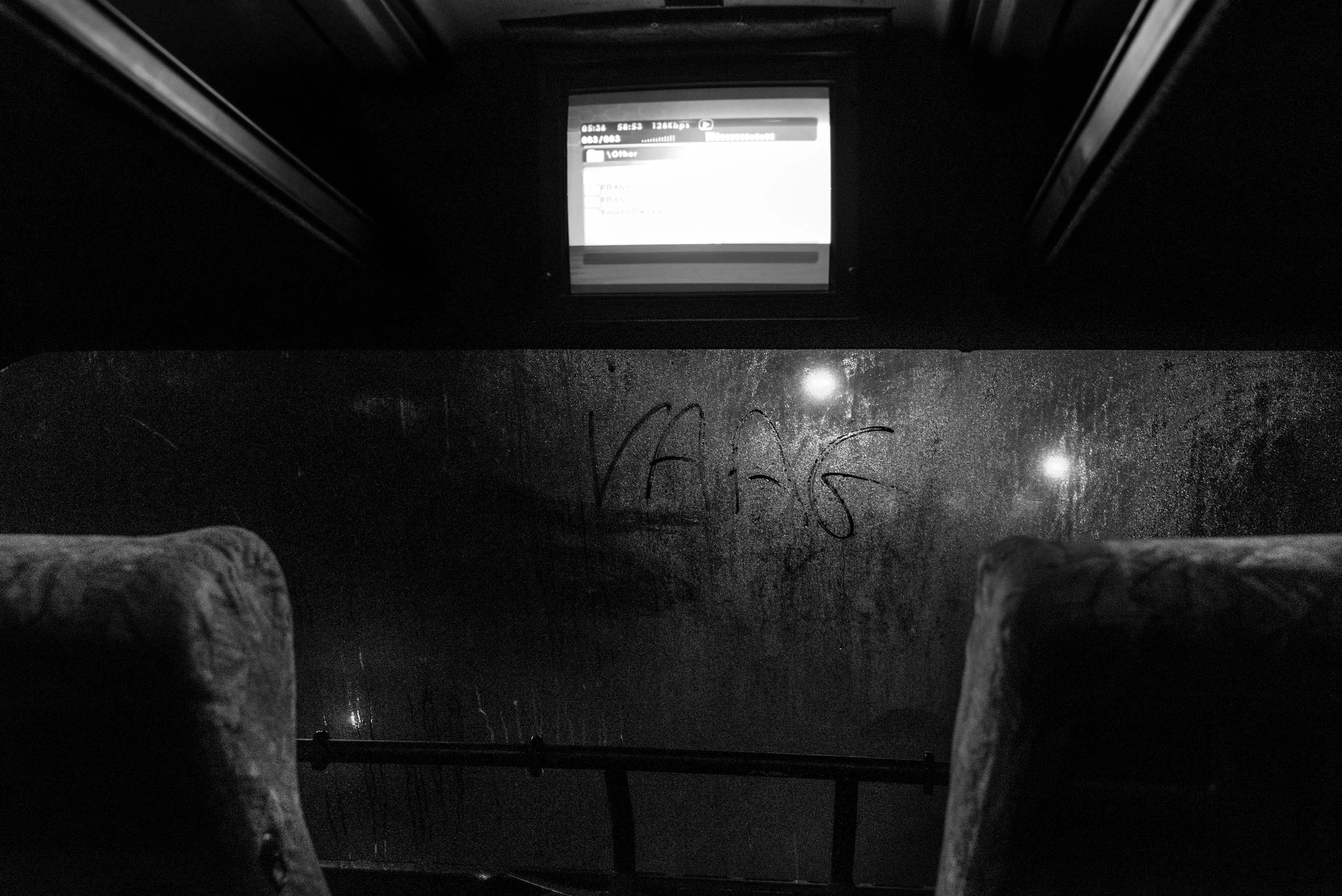 Binnenkant van een bus of trein op een regenachtige nacht, met een raambelettering die 'VAS 6' zegt en een scherm boven met informatie, beide reflecterend op de natte rand.