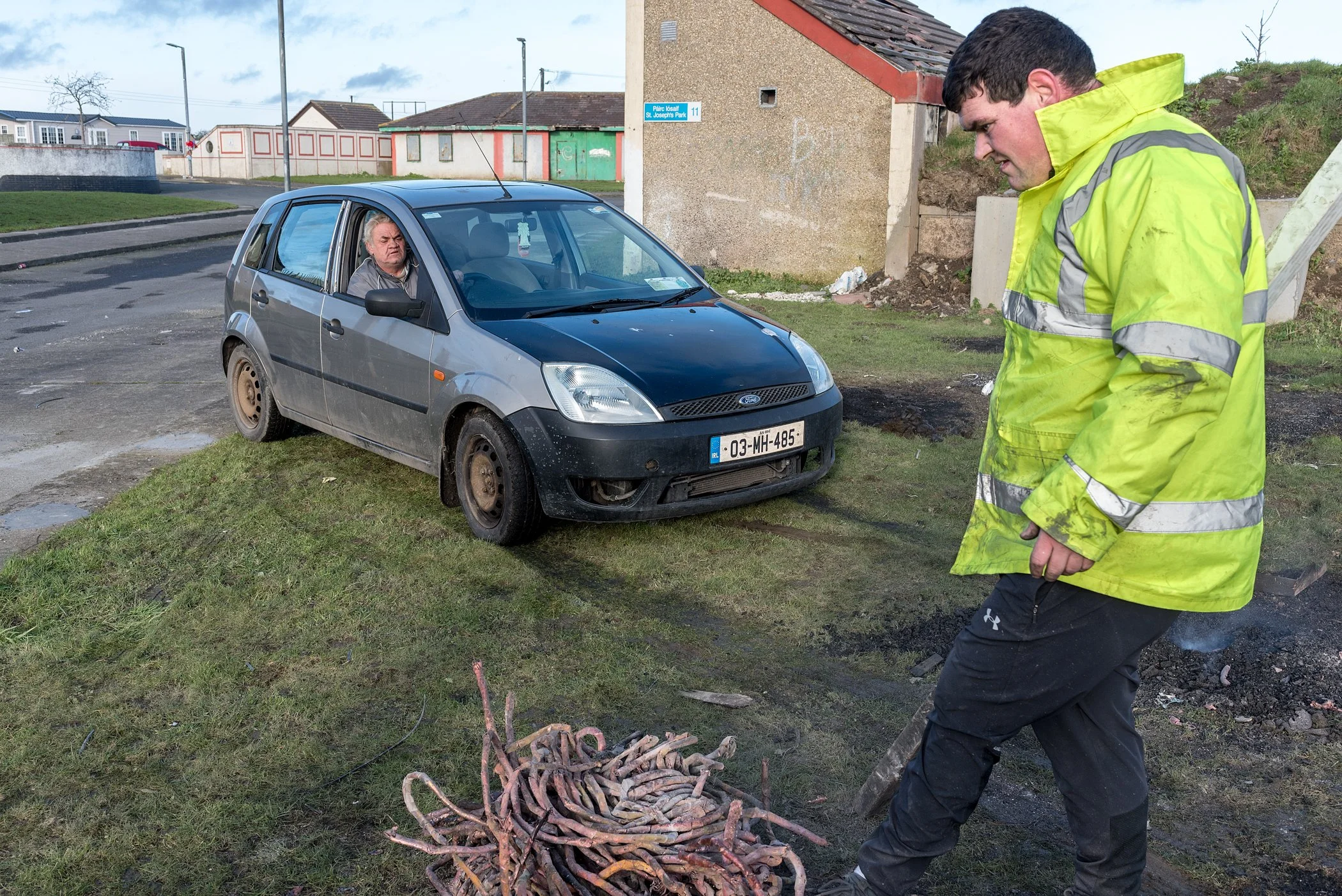 Man in een felgele veiligheidsjas die een stuk metaal vasthoudt, terwijl een geparkeerde grijze auto met een kapotte voorbumper en roestige velgen op de achtergrond staat. Er ligt roet en rook op de grond, mogelijk door een brand.