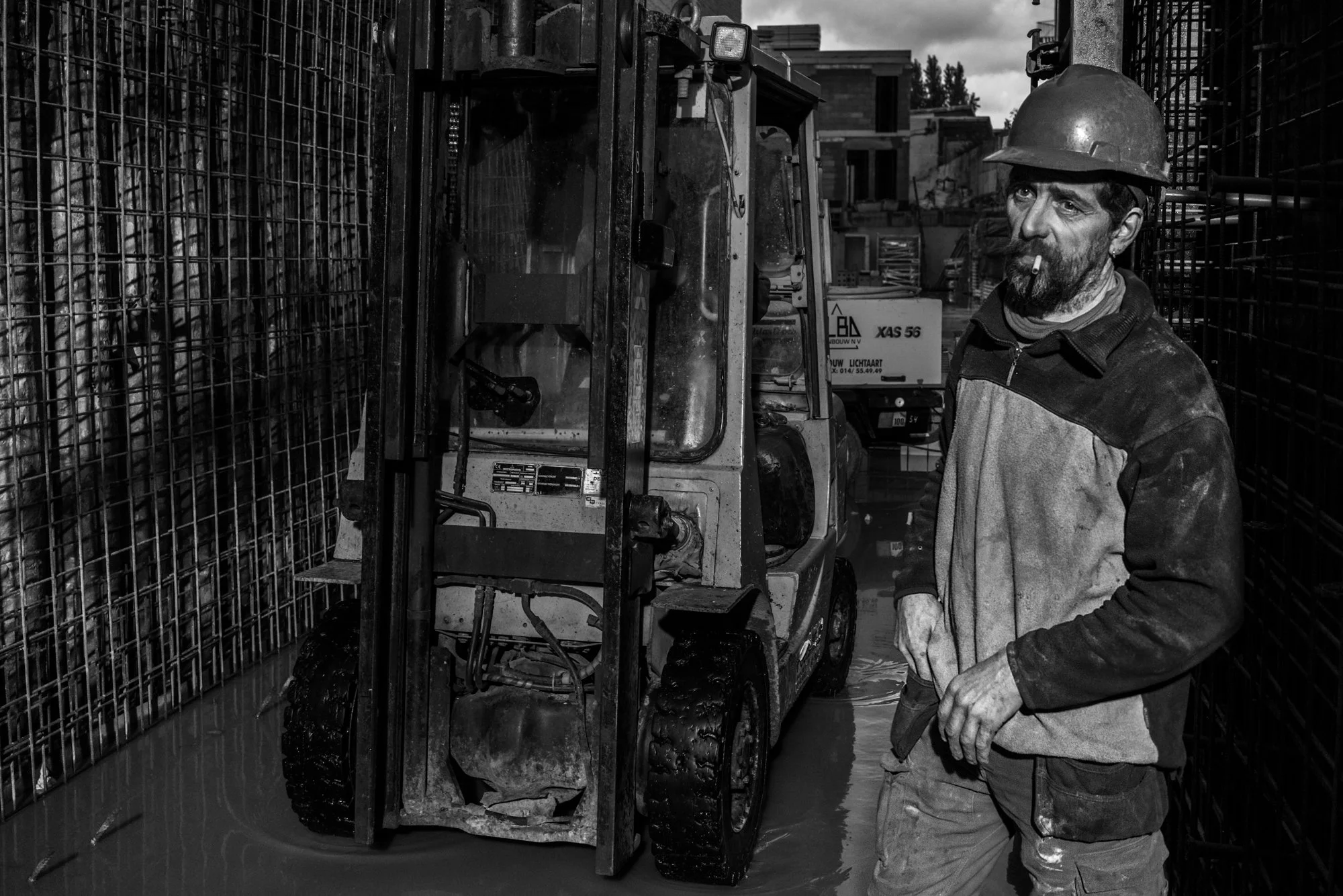 Man met bouwhelm en sigaret naast een zware machine op een bouwplaats, omringd door metalen hekwerken, in zwart-wit.