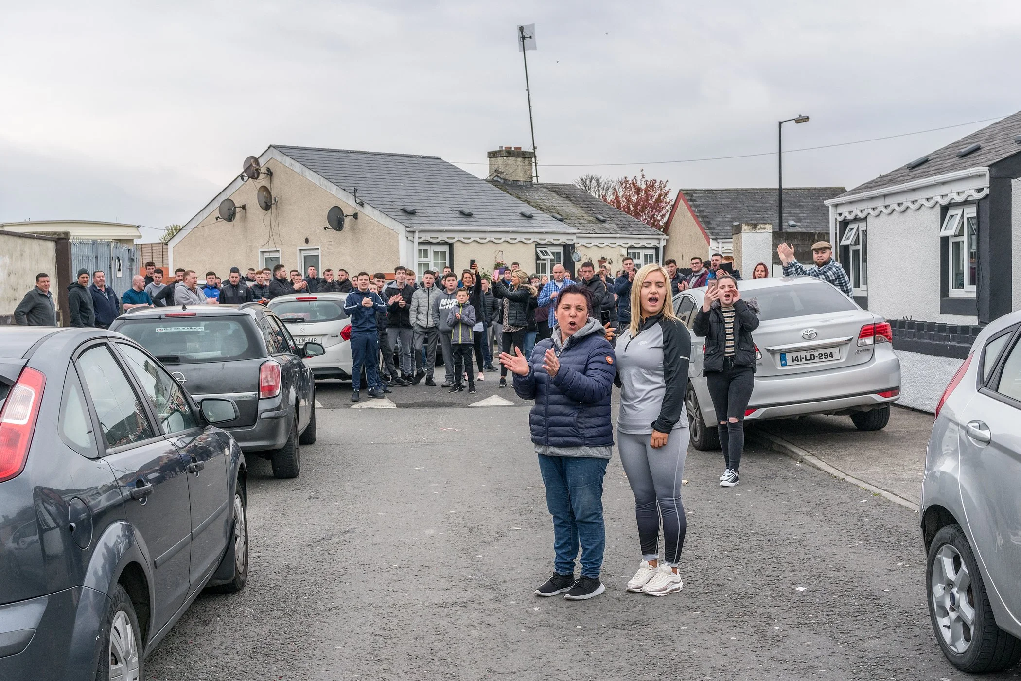 Verschillende mensen staan op een parkeerplaats waaronder een vrouw en een jongedame die fronzen en aan het praten zijn. Er is een grote groep mensen achter hen die toekijkt, sommige applaudisseren of zwaaien.