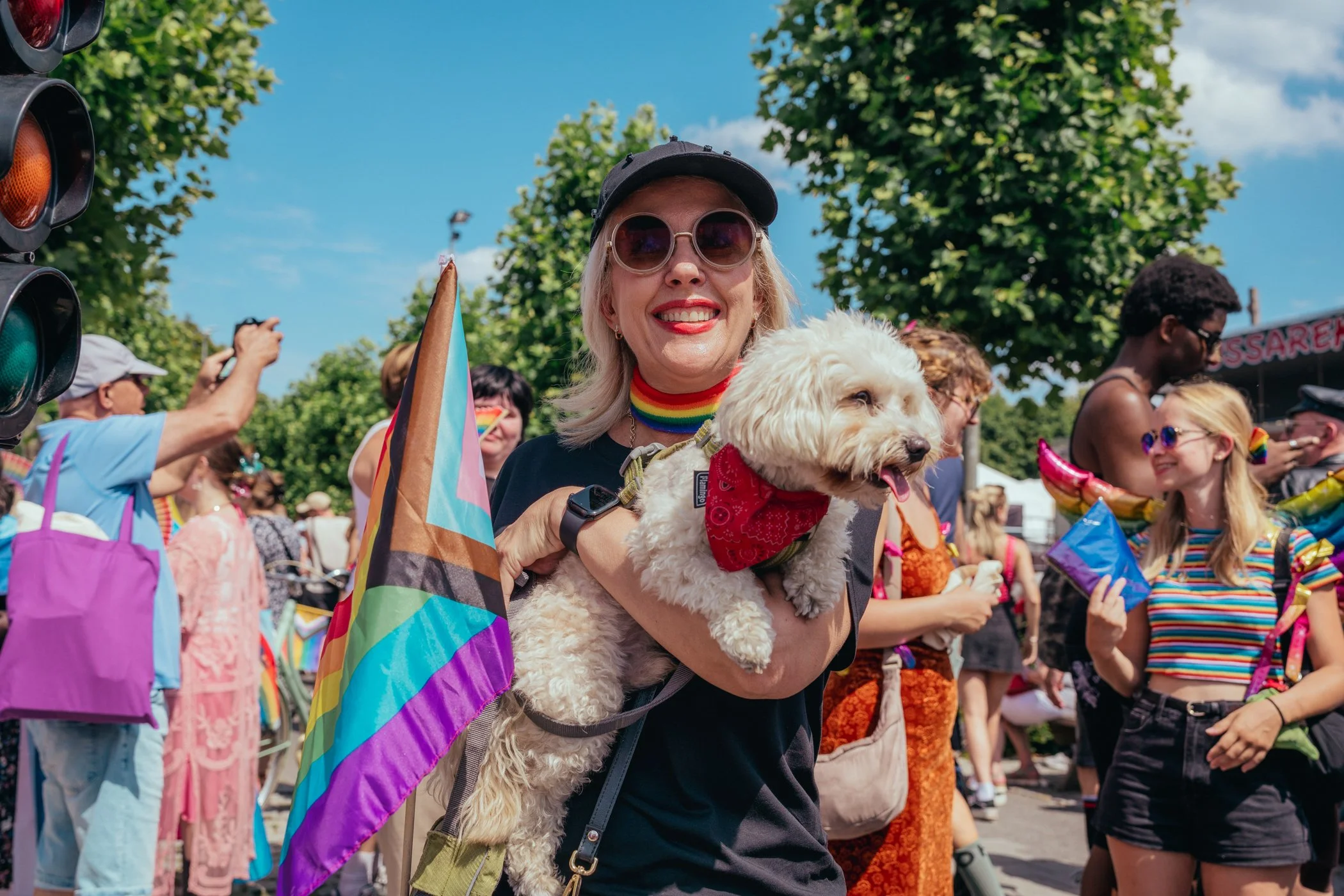 Vrouw met glimlach, sunglasses en rainbow-kraag die een hond vasthoudt, omgeven door een kleurrijk menigte tijdens een LGBTQ+-evenement.