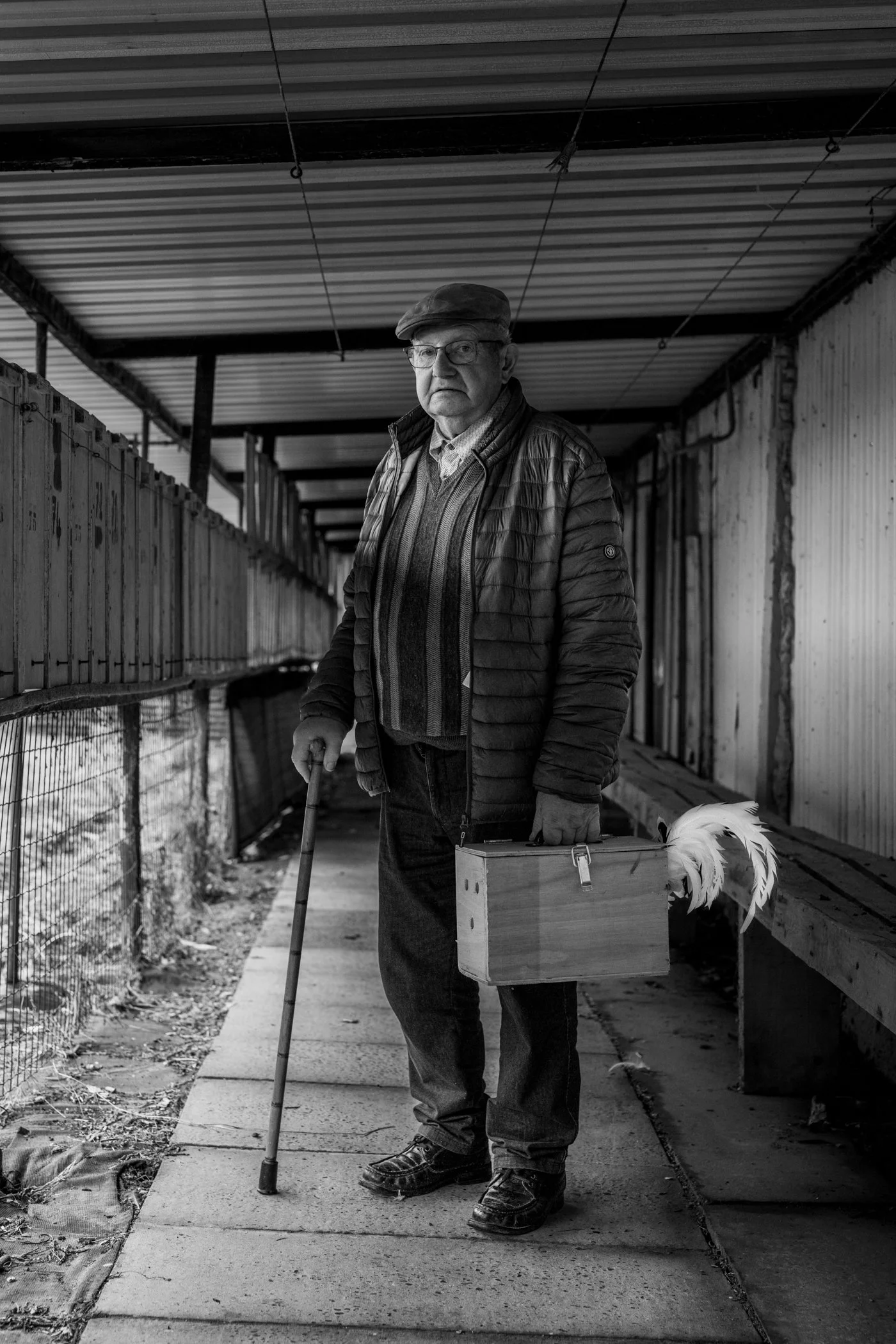 Een oudere man met een wandelstok, gekleed in een jas en een flat cap, staat op een pad binnen een soort kooi of omheining, met een houten kooitje in zijn hand.