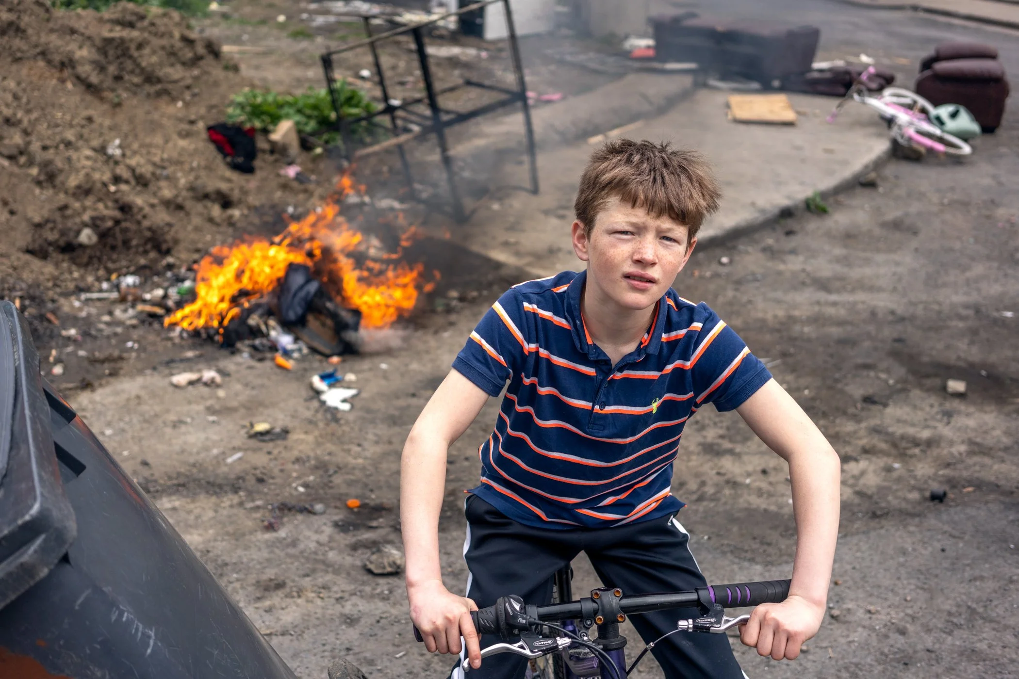 Een jonge jongen met een fiets voor zich, uitkijkend met een verwarde uitdrukking, naast een brandende vuilnisbak en een campingbed.