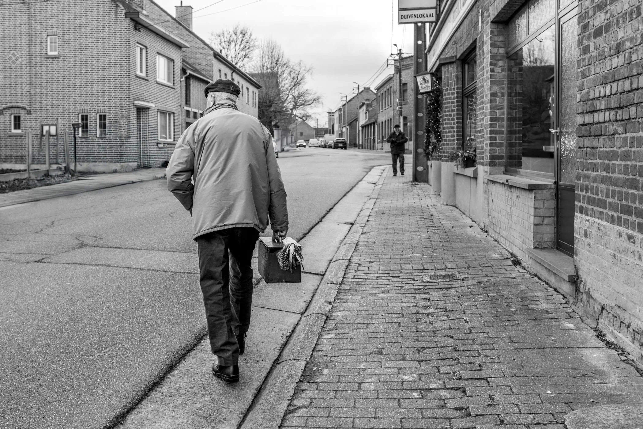 Een man loopt met een kistje op straat, op een rustige wijk met stedelijke gebouwen en een paar mensen op de achtergrond, in zwart-wit.