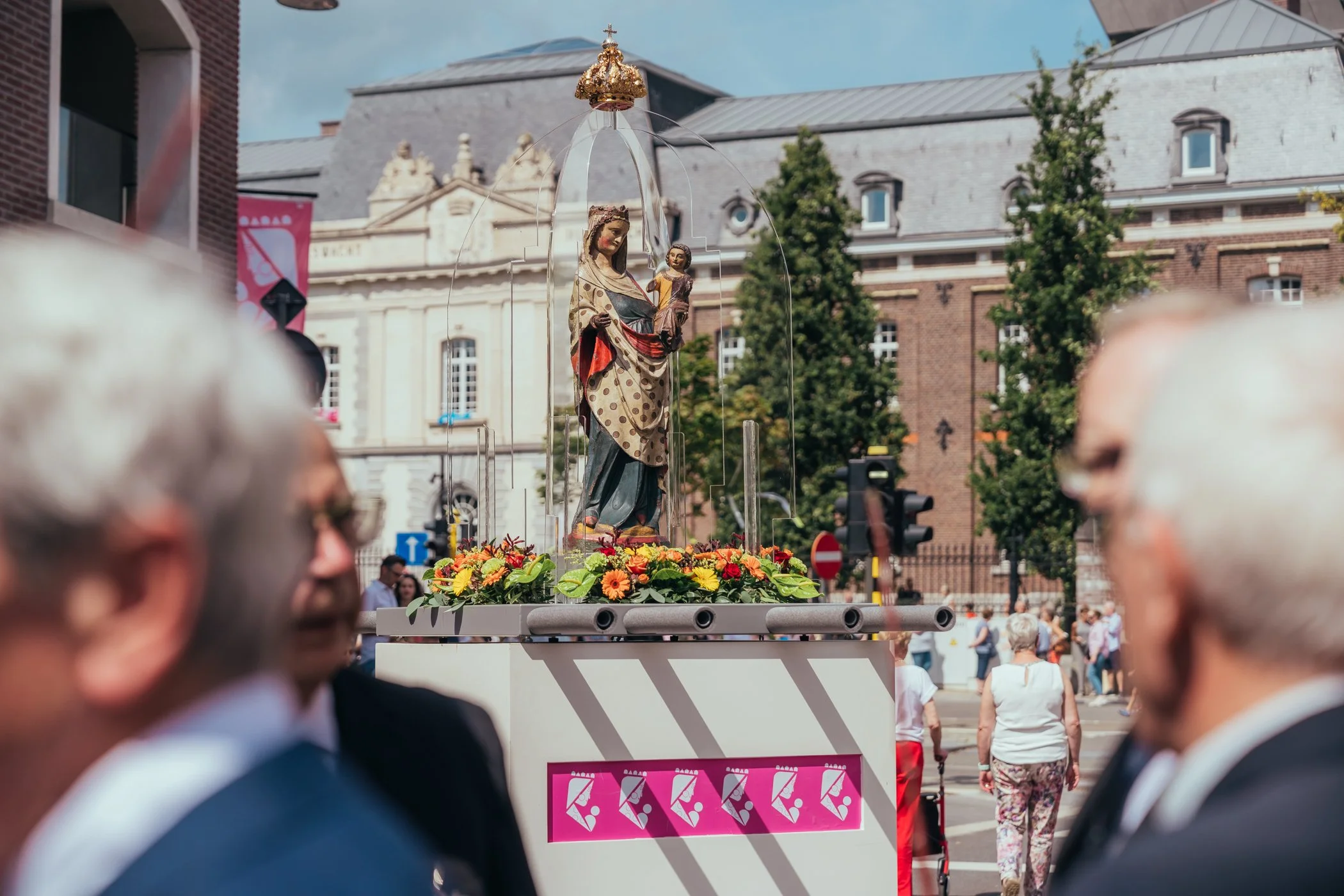 Een straatbeeld met een processie waarbij een standbeeld van de Maagd Maria met kind op een houten platform staat, versierd met bloemen, omringd door mensen.