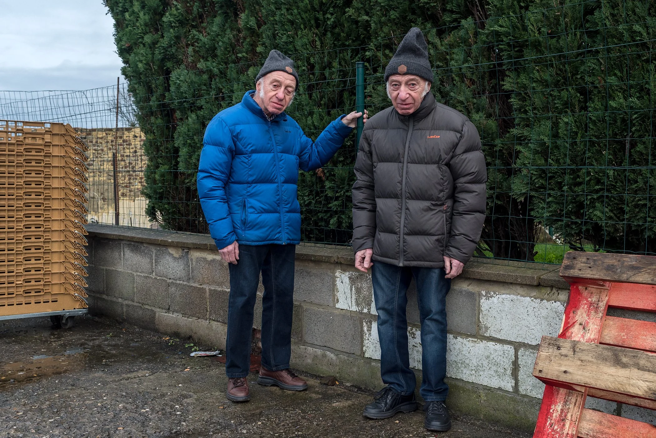 Twee oudere mannen in jassen en mutsen staan buiten tegen een hek en een muur, met een bos achter hen.