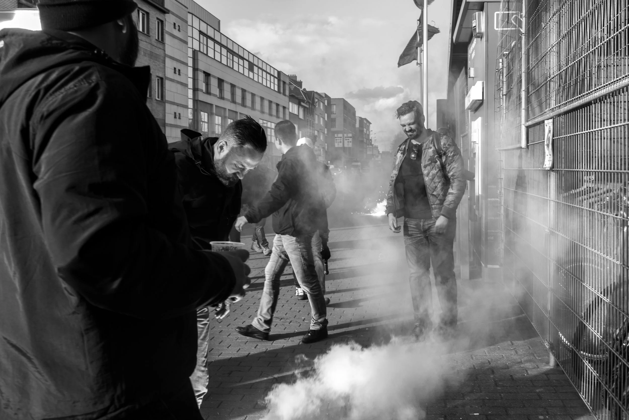 Mensen op straat met rook en een brandend object, schemerige omgeving met gebouwen en mensen die bezig lijken te zijn met protest of rellen.