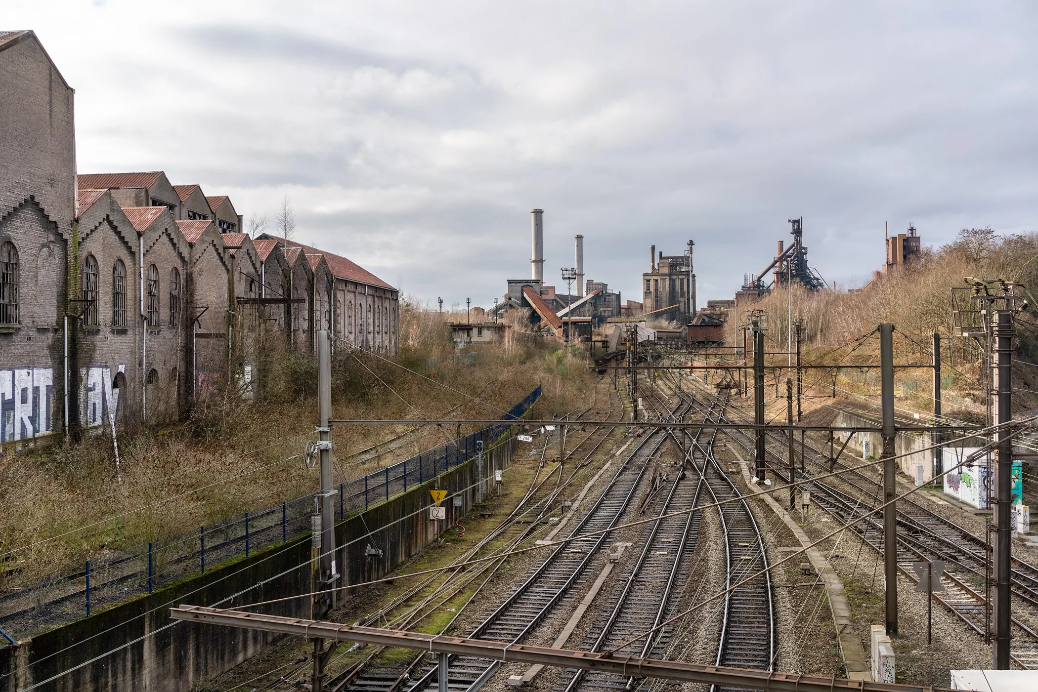 De afbeelding toont een spoorwegnetwerk met meerdere sporen en elektrische lijnen, omgeven door industrieel landschap met oude fabrieken op de achtergrond en graffiti op de muur aan de linkerkant.