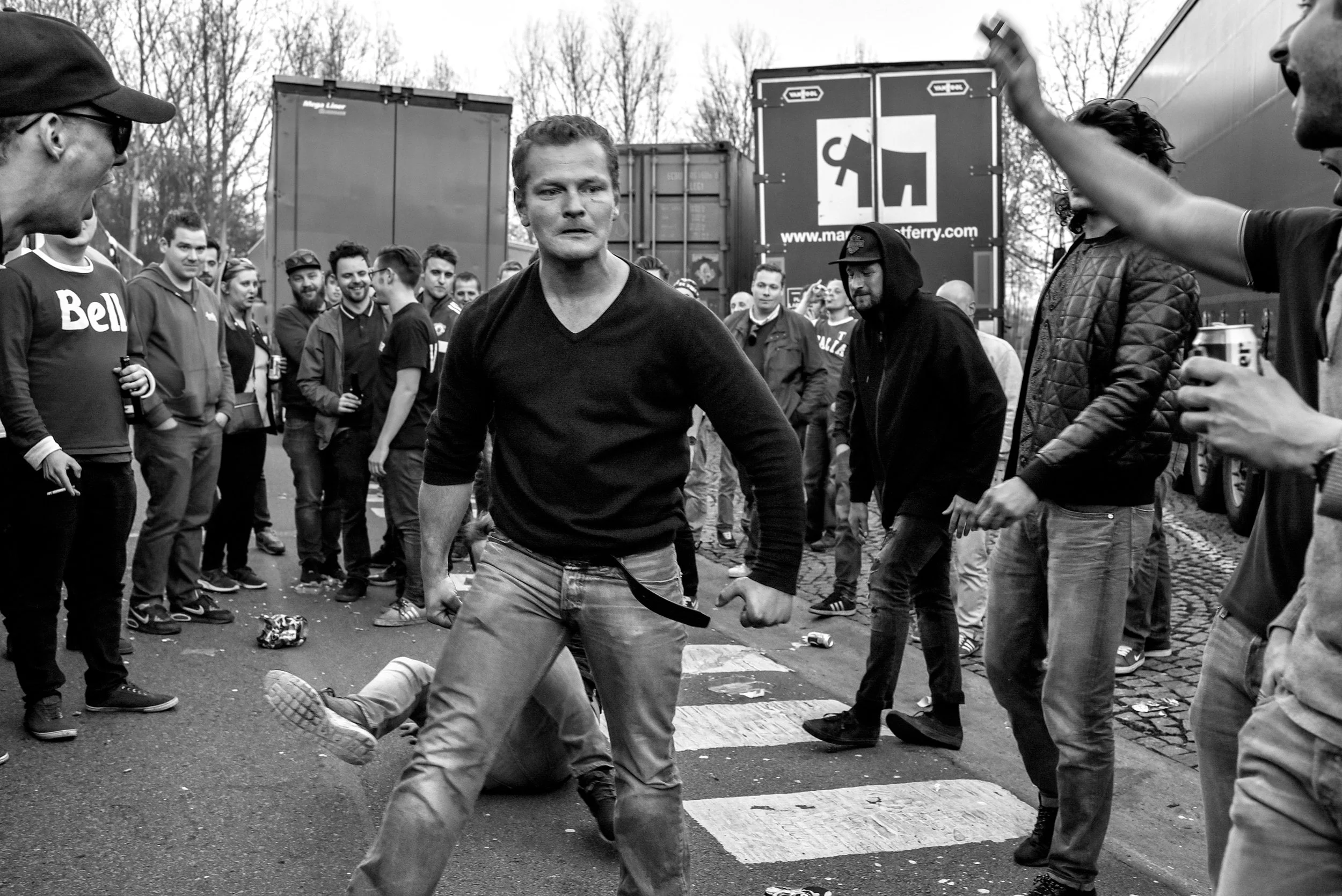 Een zwart-wit foto van een straat, met een man in het midden die lijkt te dansen of te bewegen, omringd door een groep mensen die kijken en some drinken vasthouden.