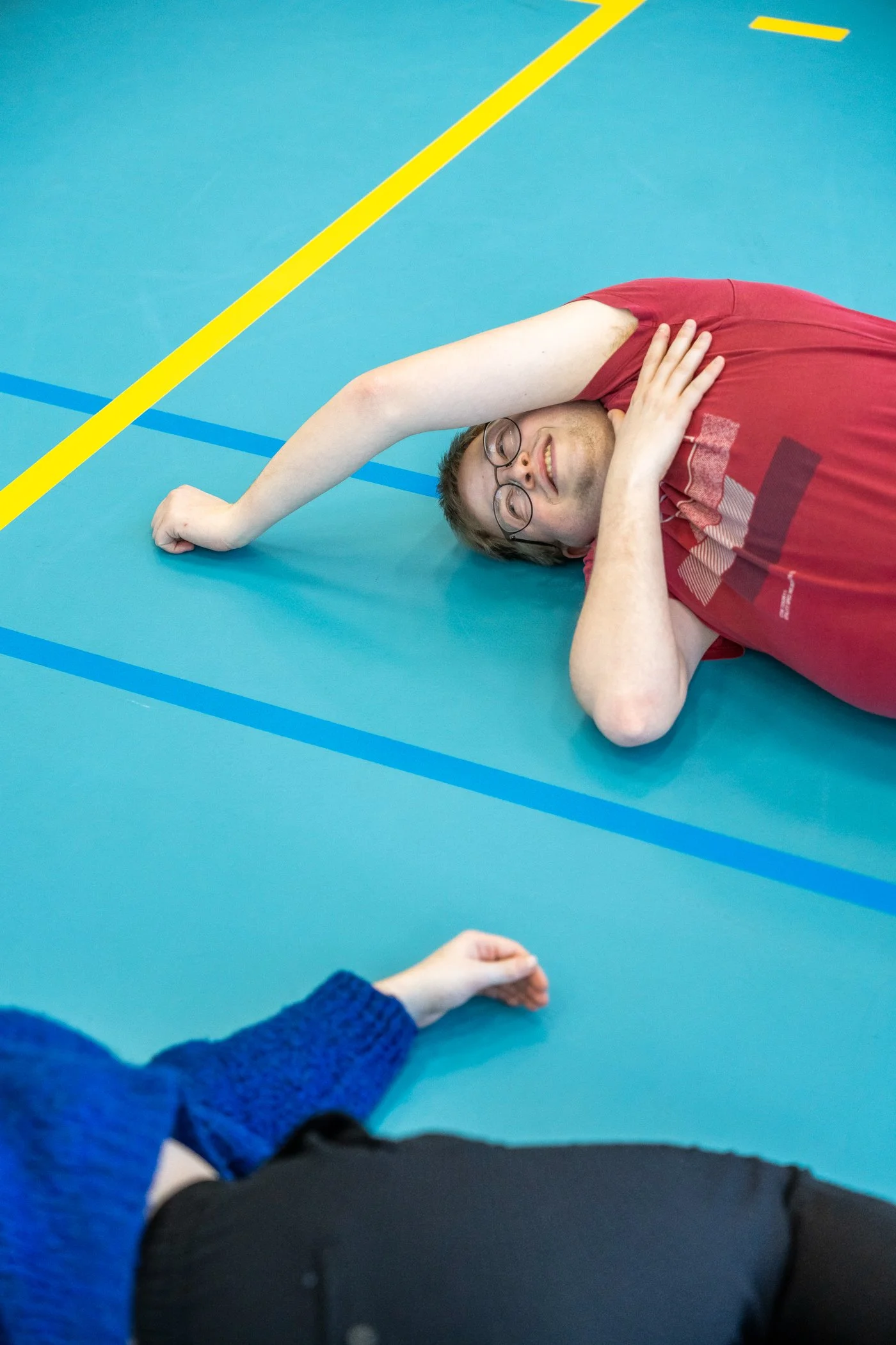 Man ligt op de vloer met een arm over zijn gezicht, terwijl een ander persoon in een blauwe trui naast hem ligt op de vloer, de setting lijkt een sporthal met blauwe vloeren en gele lijnen.