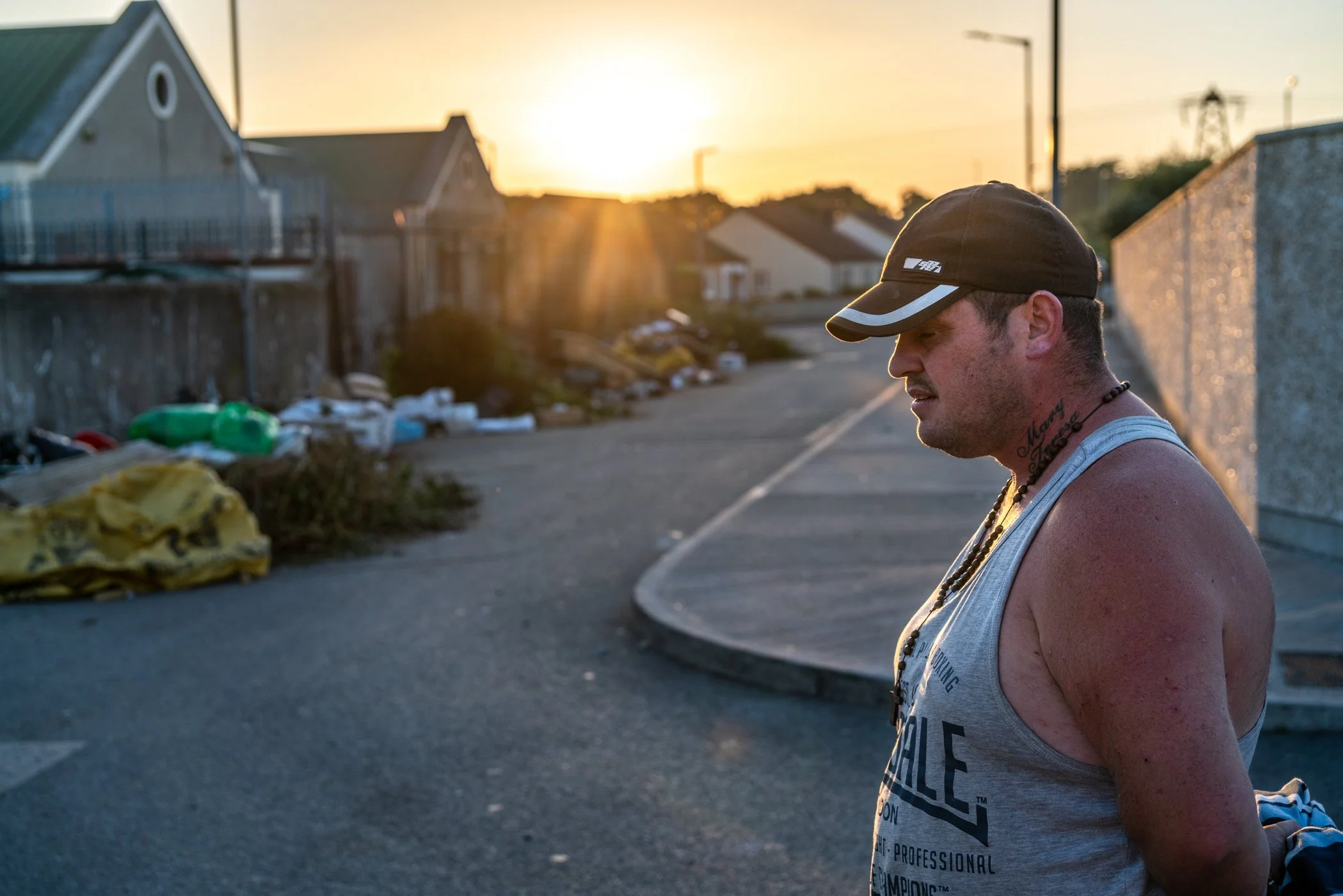 Een man staat op een straat tijdens de zonsondergang, hij draagt een grijze tanktop, een zwarte pet en een ketting, en kijkt naar beneden.