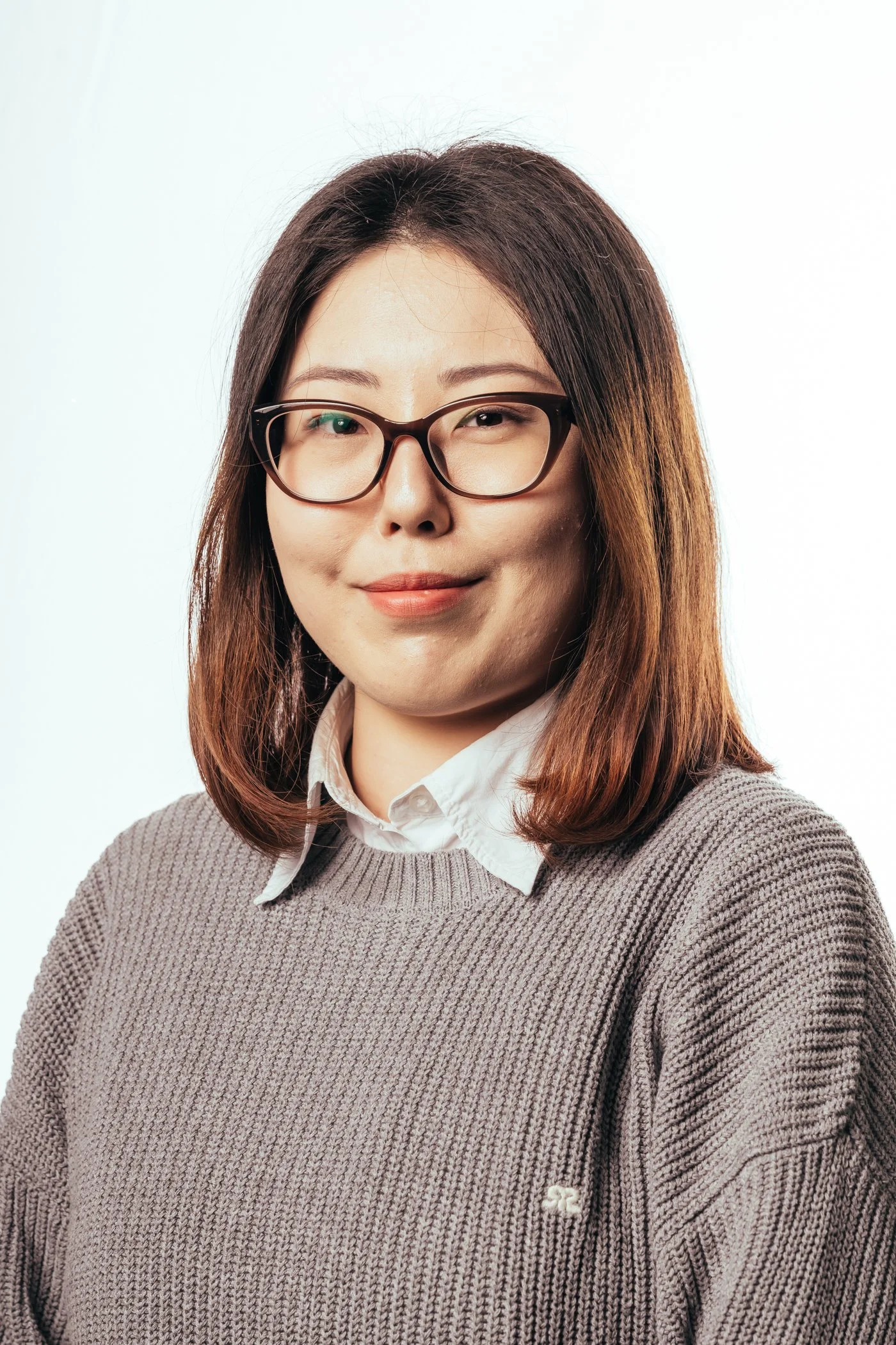 Portret van een vrouw met bruin haar, bril, witte blouse en grijze trui tegen een witte achtergrond.