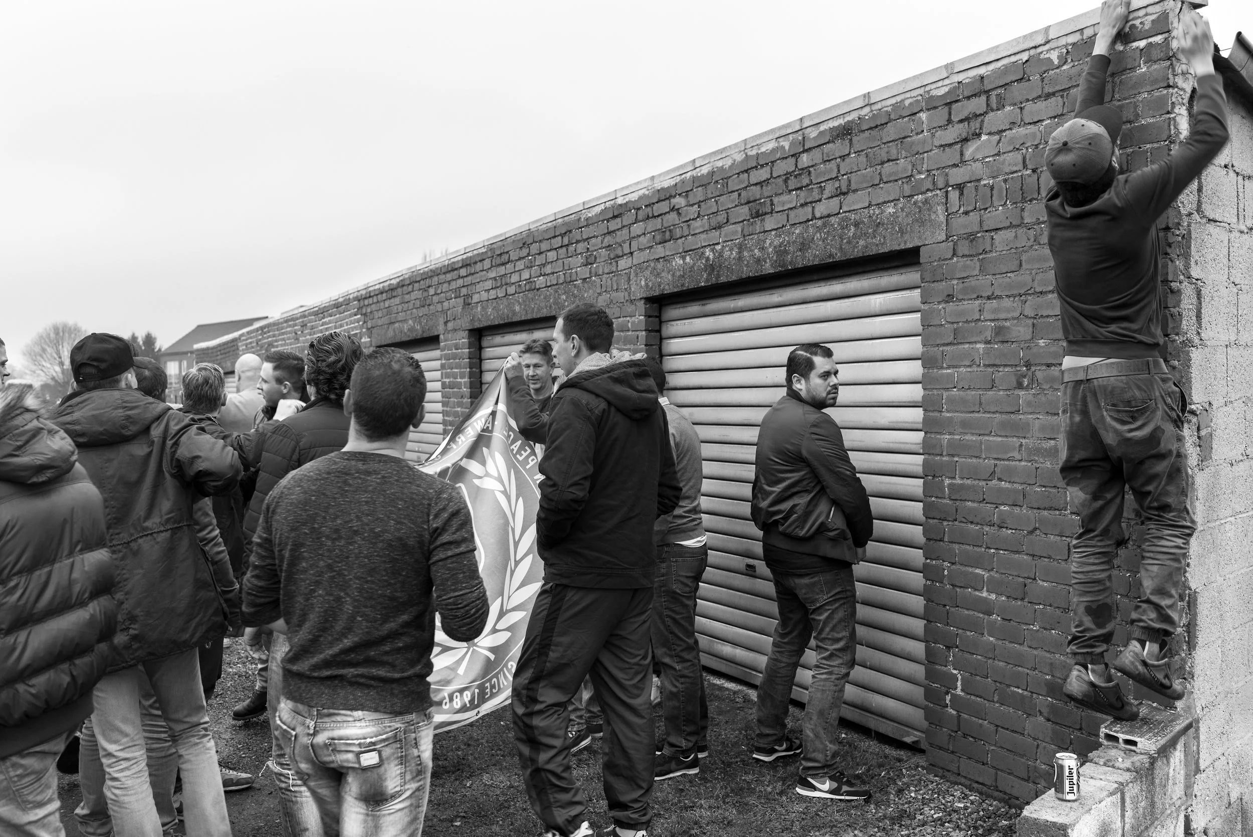 Mensen bij een protest of demonstratie rondom een gesloten garage deur, met één persoon die aan de muur hangt en een persoon die een vlag vasthoudt.