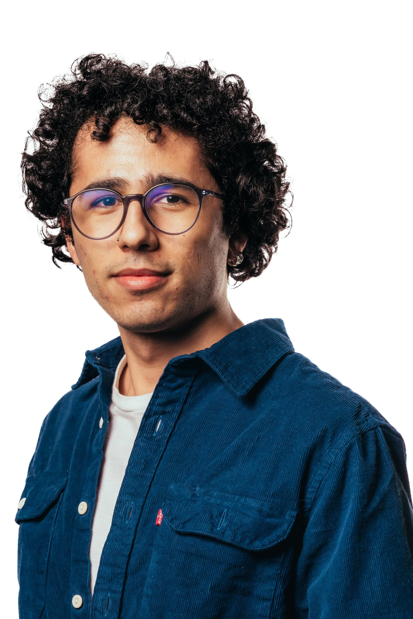 Portret van een jonge man met krullend haar, bril, lichte huid en casual kleding, lachend op een witte achtergrond.