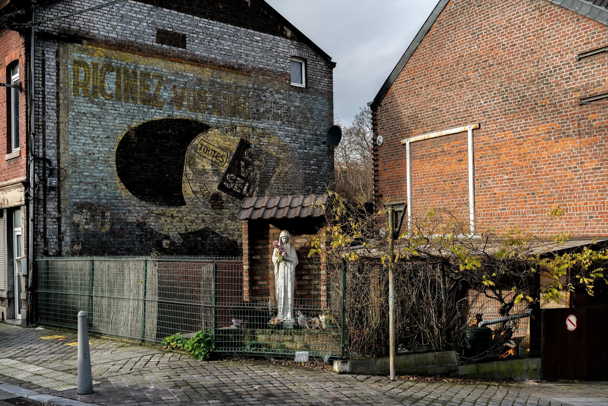 Een straat met een muurschildering en een religieuze beeldhouwwerk van een heilige. De muur heeft een grote oude reclame die lijkt op een gebied met de tekst 'RIGINEZ VIS' en een afbeelding van een bidon. De straat heeft een stenen stoep en een hek r
