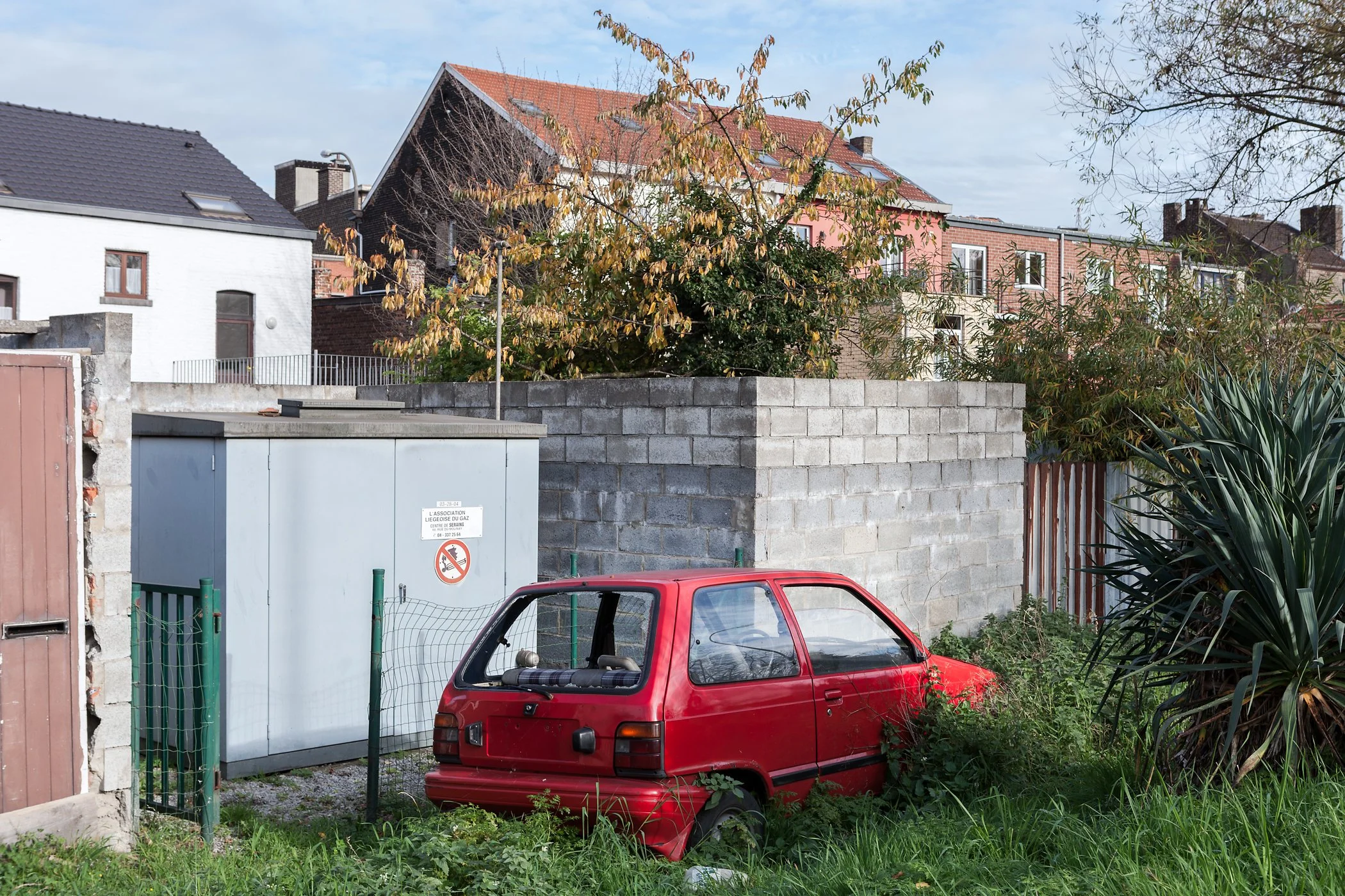 Een oud rode compacte auto staat op een grasveld, omgeven door groen en struiken, naast een grijze klein gebouw en een stenen muur, met in de achtergrond huizen met verschillende gevels en schuine daken.