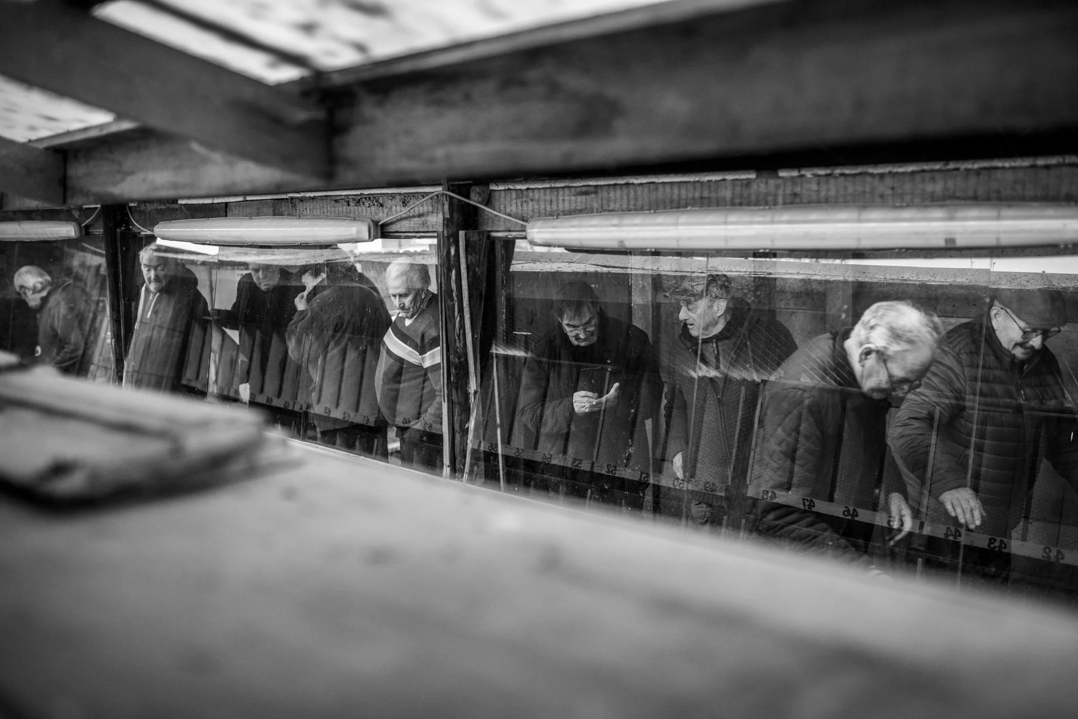 Zes mensen staan in een rij achter een glazen scheidingswand, gekleed in donkere jasjes en hoeden, naar beneden kijkend op een station of platform in zwart-wit.