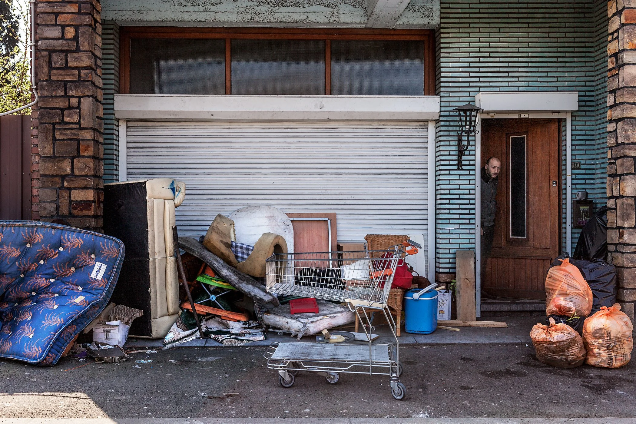 Een huis met een gesloten rolluik en een deur, omgeven door diverse afval en spullen zoals een matras, bureaustoelen, een winkelwagen, afvalzakken en andere voorwerpen, op de zomerse straat.