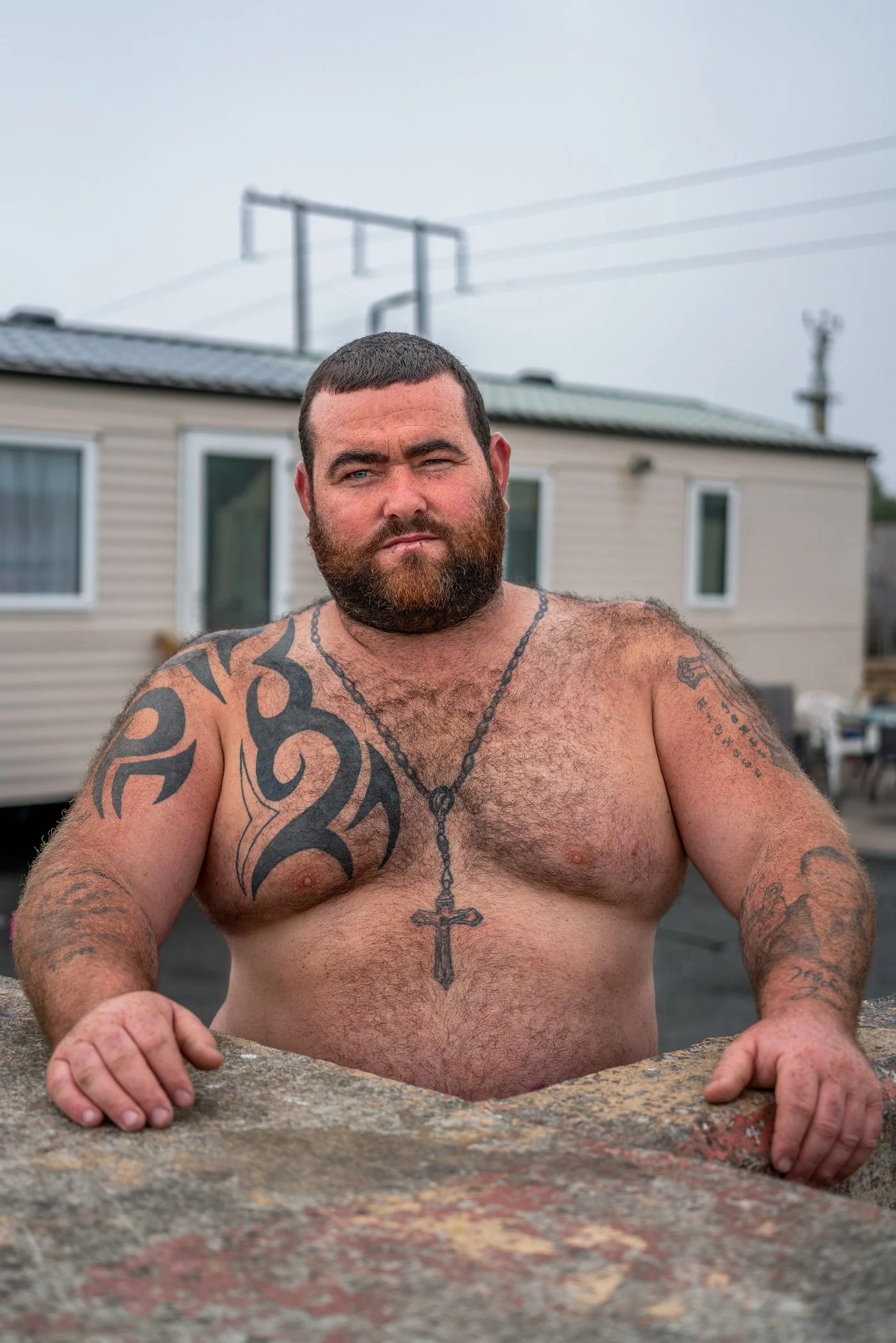 Een shirtloze man met een volle baard en tatoeages op beide armen, die in een bad hangt, met een huis en een elektrische draad in de achtergrond.