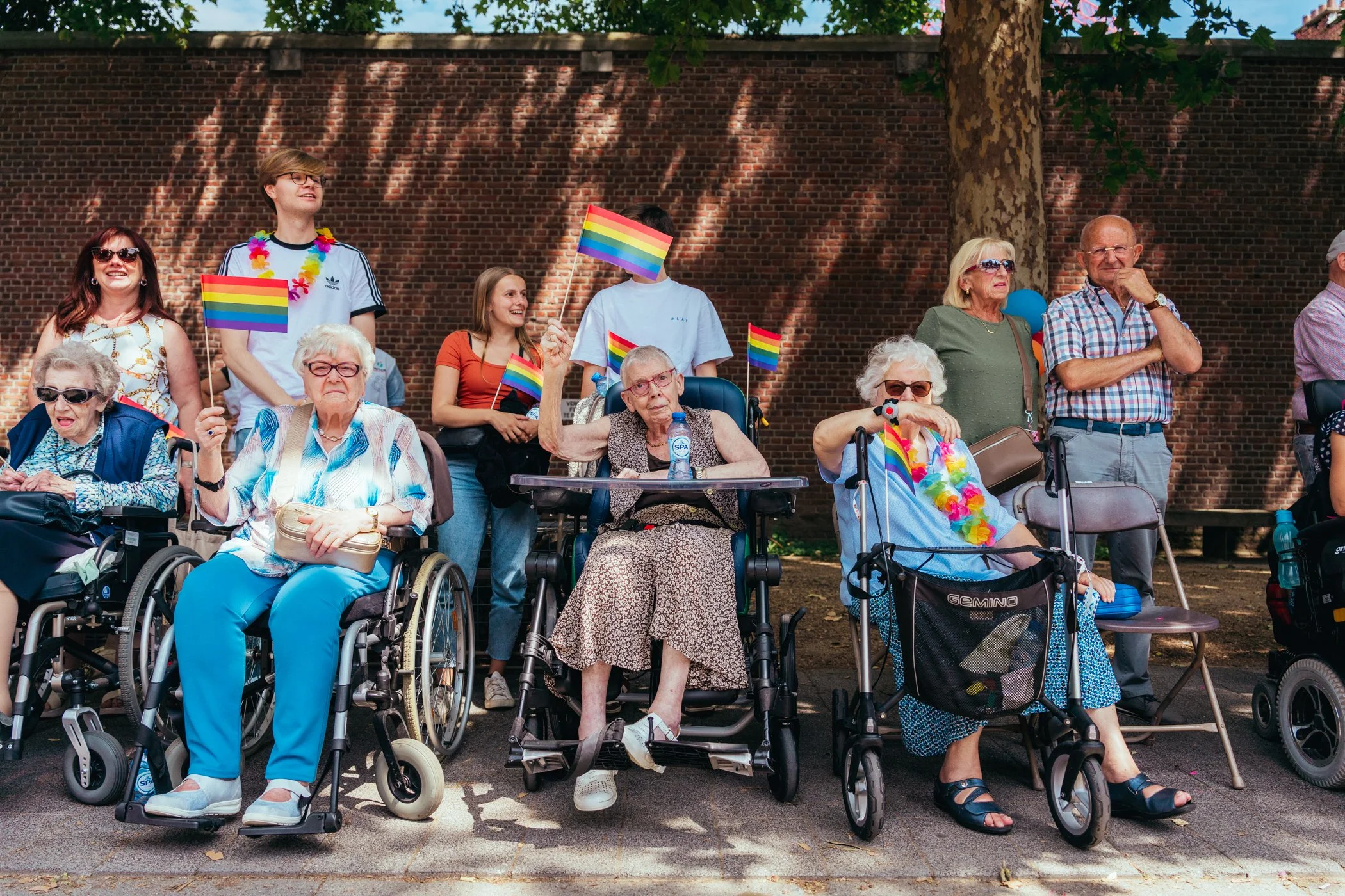 Groep ouderen en jonge mensen poseert tijdens een Pride-evenement met regenboogvlaggen en kleurrijke accessoires, in de buitenlucht tegen een bakstenen muur.