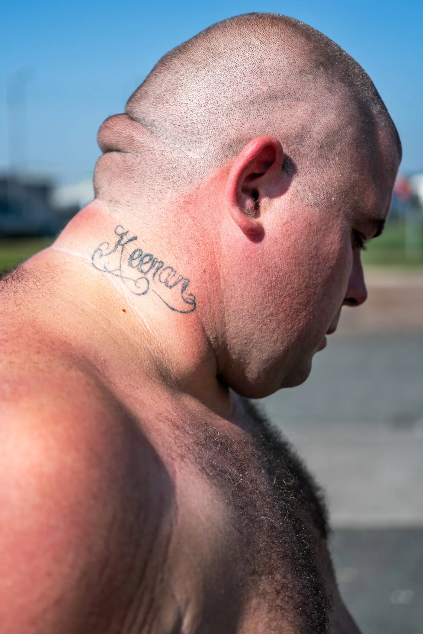 Close-up van een naakte man met een tattooschrift 'Keenon' op zijn nek, donker haar en een korte kapsel, buiten op een zonnige dag.