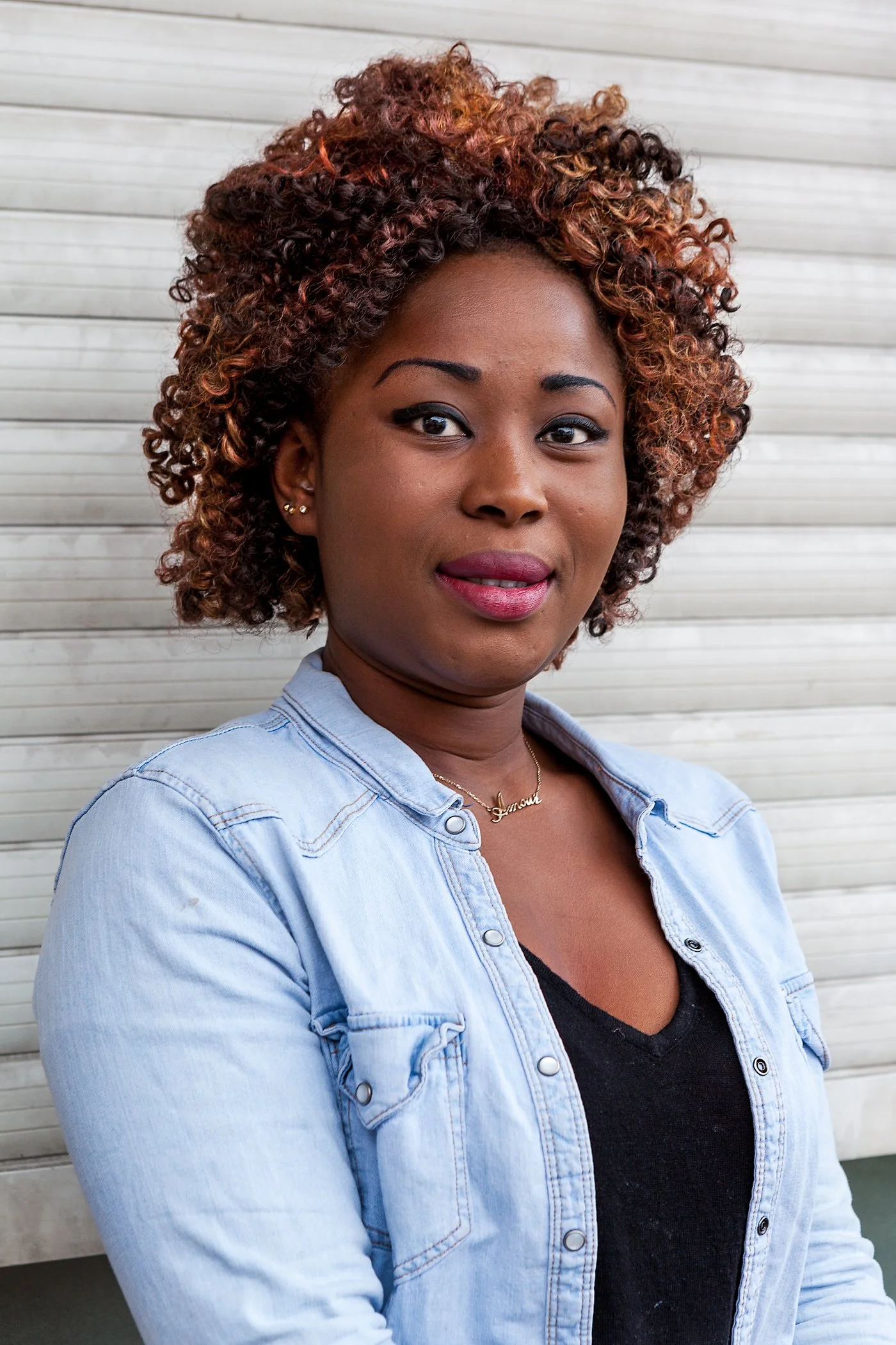 Portret van een vrouw met gekruld, kleurrijk haar, zilveren oorbellen en een halsketting met naam, gekleed in een denim jas en een zwart shirt, staand voor een witte houten muur.