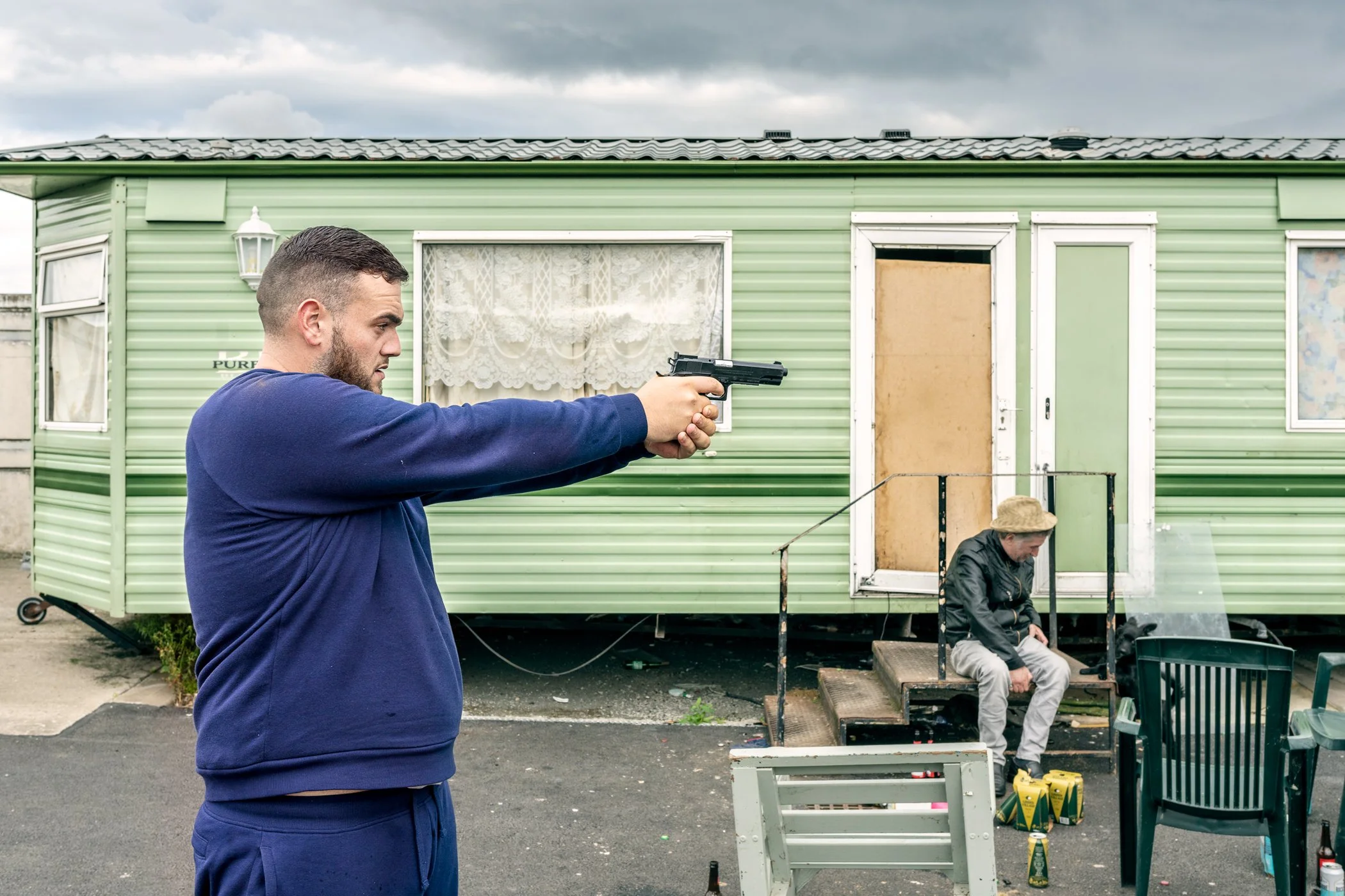 Man richt een pistool op een mobiele woning, terwijl een vrouw op de veranda zit.