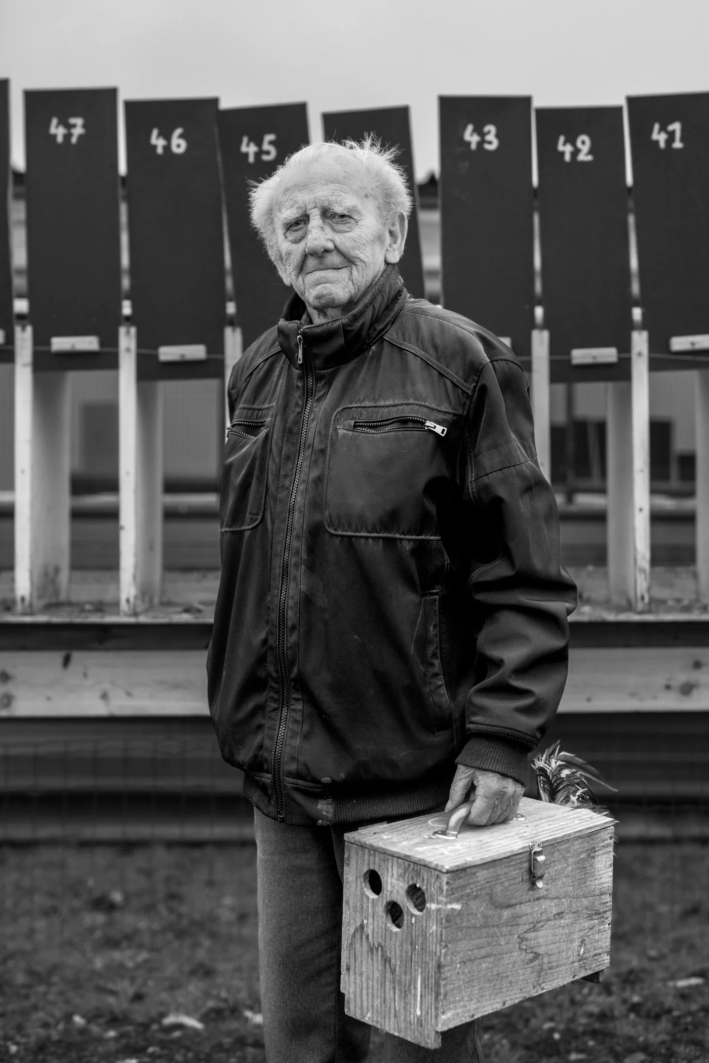 Een oude man met wit haar draagt een leren jas en houdt een houten vogelhuis met een touw vast. Achter hem staan stemkastjes met nummerplaten en wandombonden.