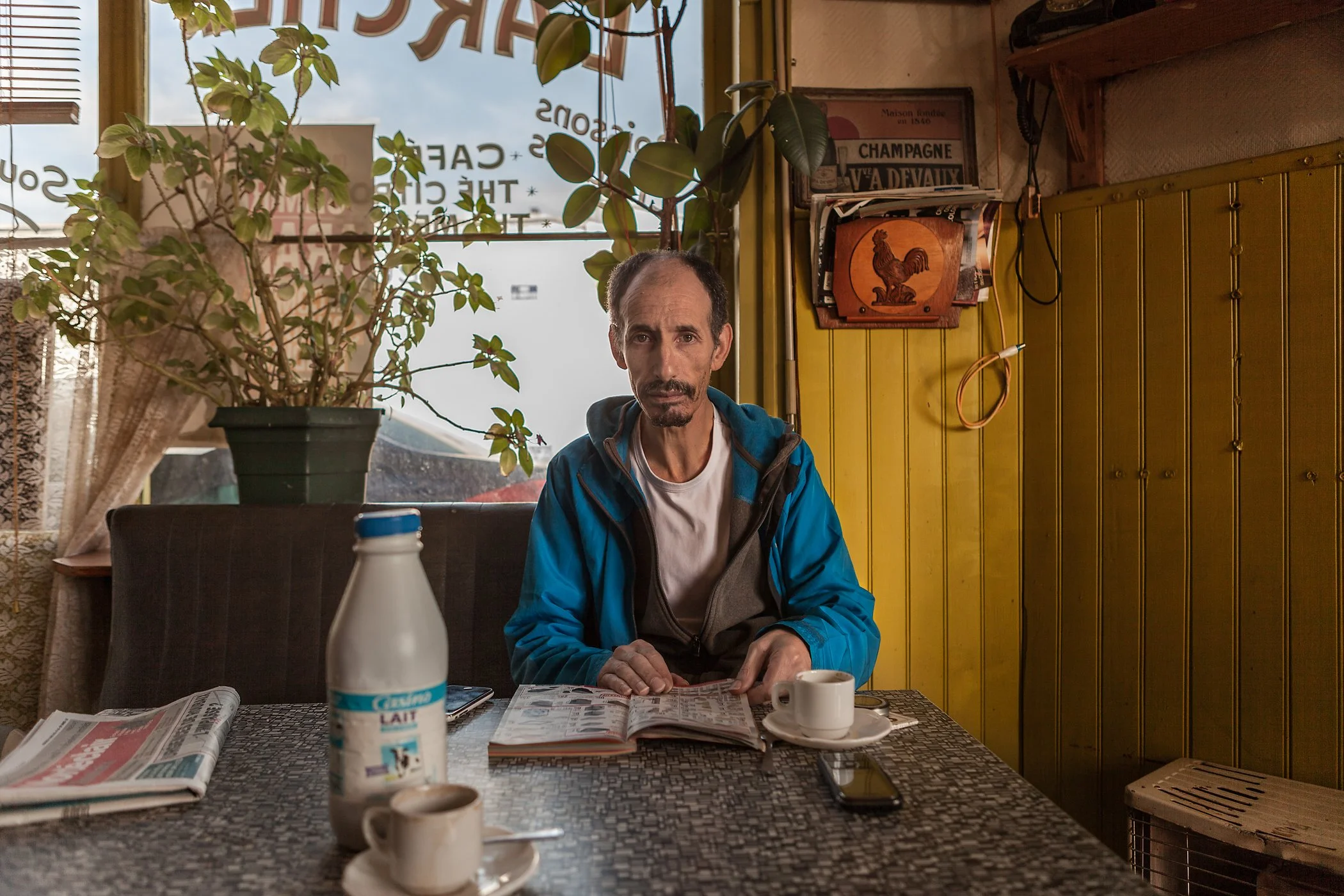 Man zit aan een tafel in een café, met een krant, een boek, een kopje koffie, een fles melk, een telefoon, en een kleine kopje koffie. Achter hem is een grote plant en een geel houten muur met decoraties.