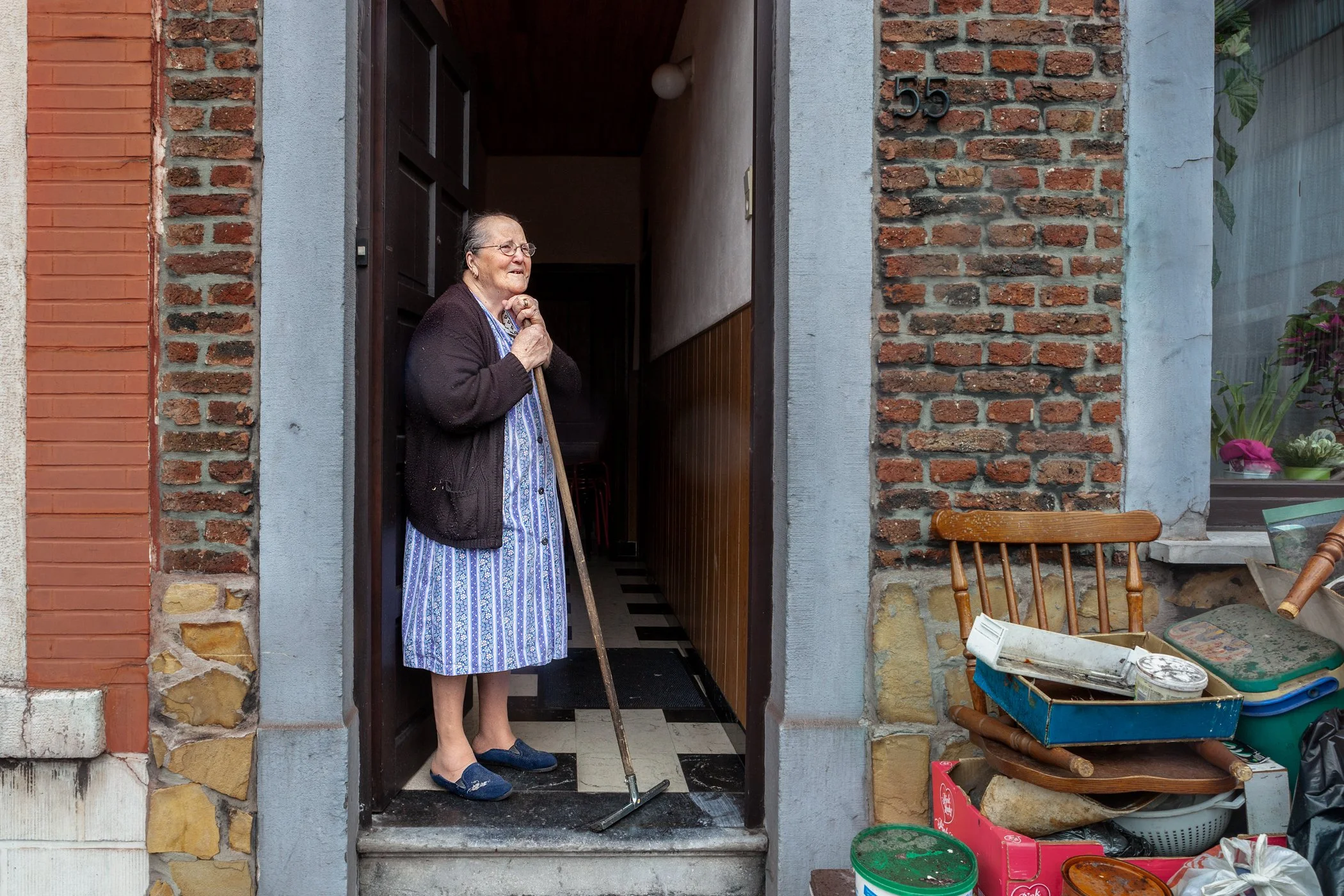 Een oudere vrouw staat in de deuropening van een huis, met een wandelstok in haar hand, gekleed in een blauwe jurk en een donkerbruine cardigan, terwijl ze uitkijkt.