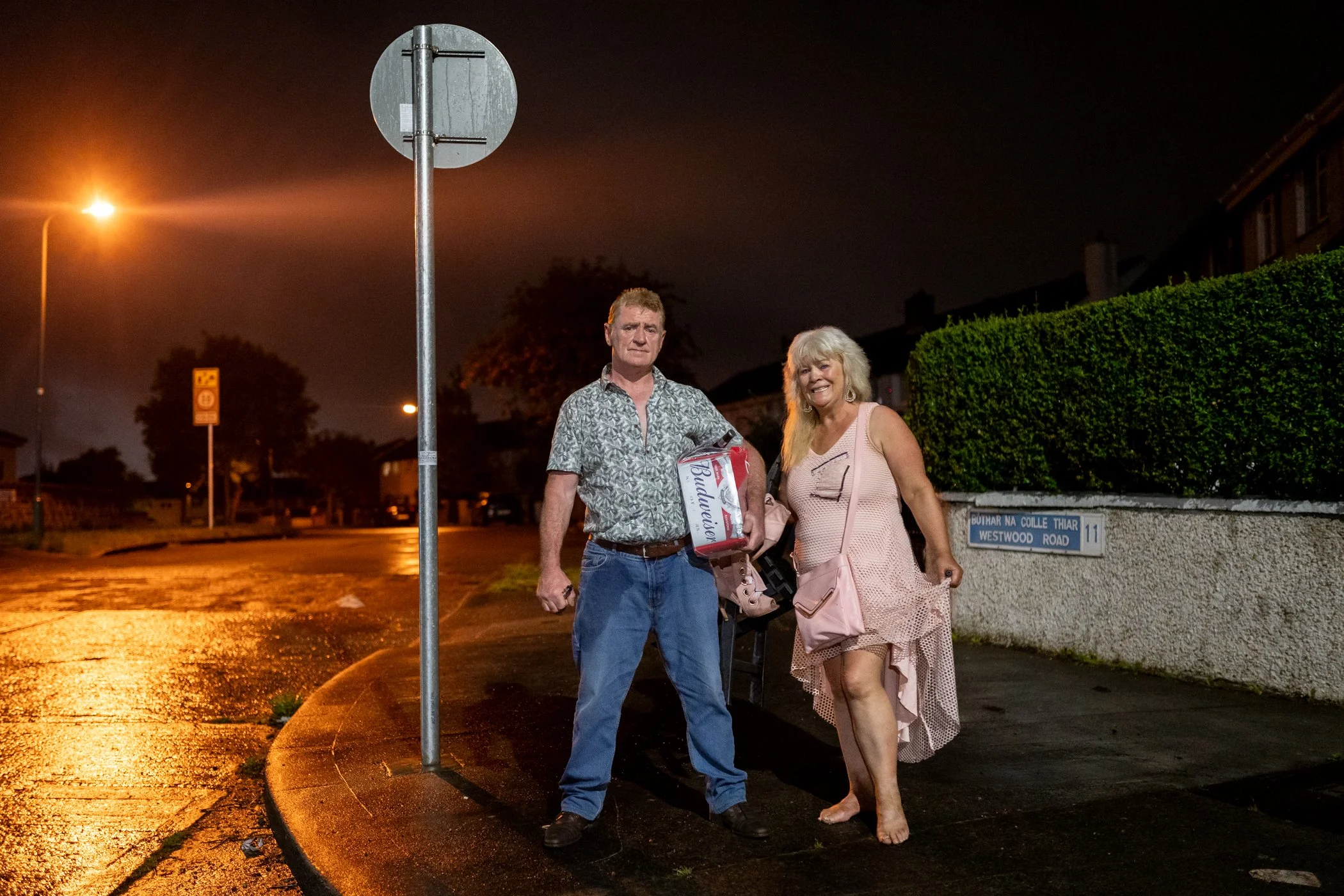 Een man en een vrouw staan samen op een natte stoep 's nachts. De man houdt een doos bier vast, de vrouw is gekleed in een roze jurk en heeft een tas. Ze lachen en poseren voor de camera.