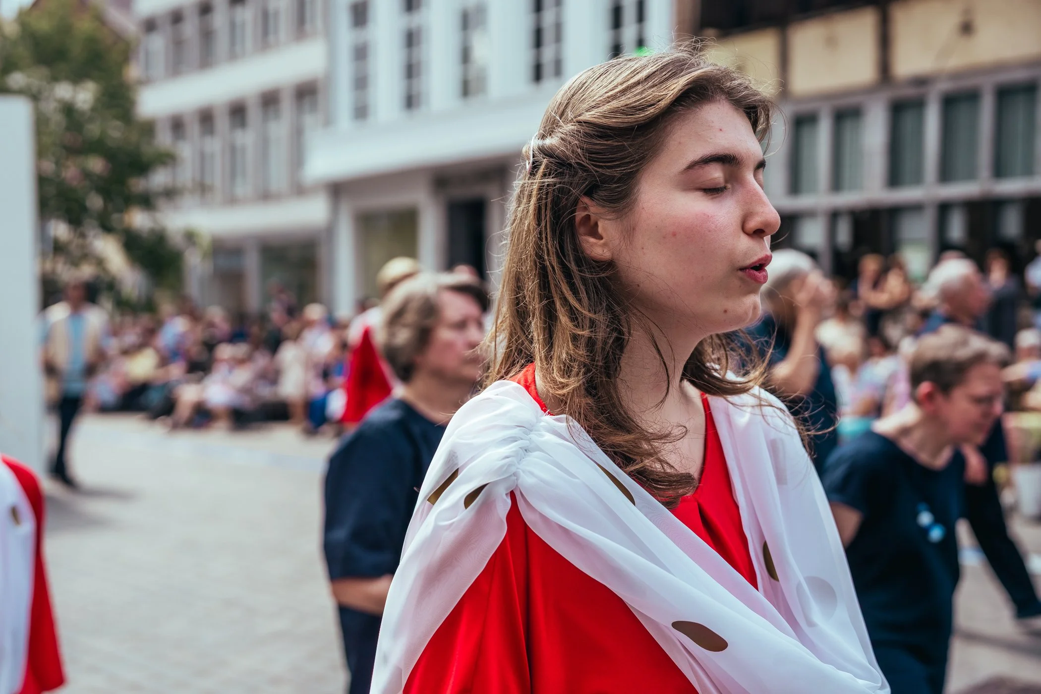 Een jonge vrouw met haar ogen dicht, gekleed in een rode jurk en een witte sjaal met goudkleurige stippen, omringd door een menigte mensen op straat tijdens een evenement.