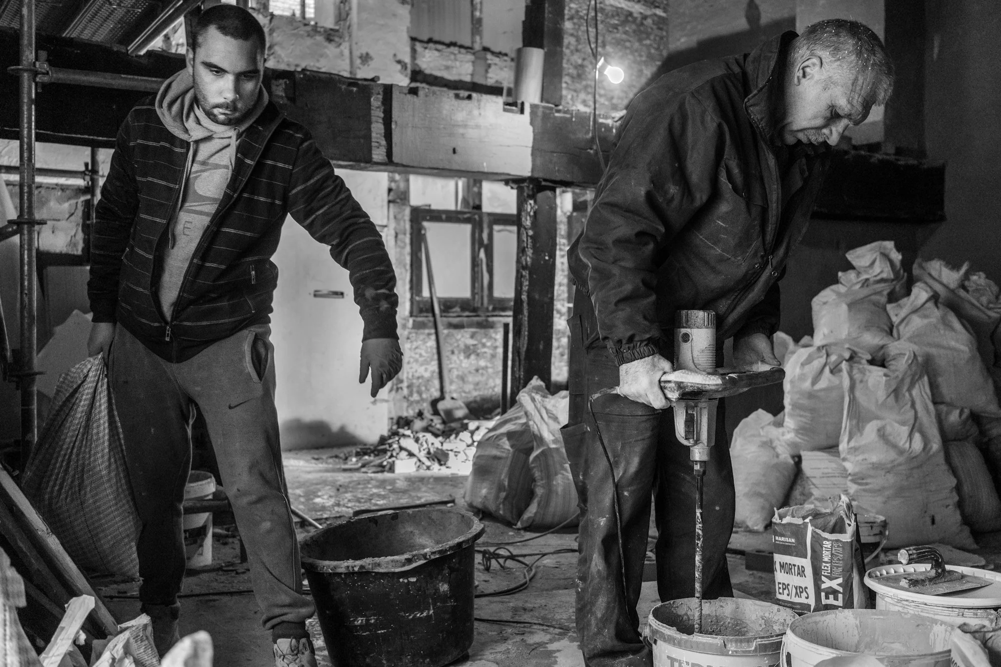 Twee mannen werken in een bouwplaats, één met een boormachine en de ander met een emmer, omgeven door bouwmaterialen en afval.