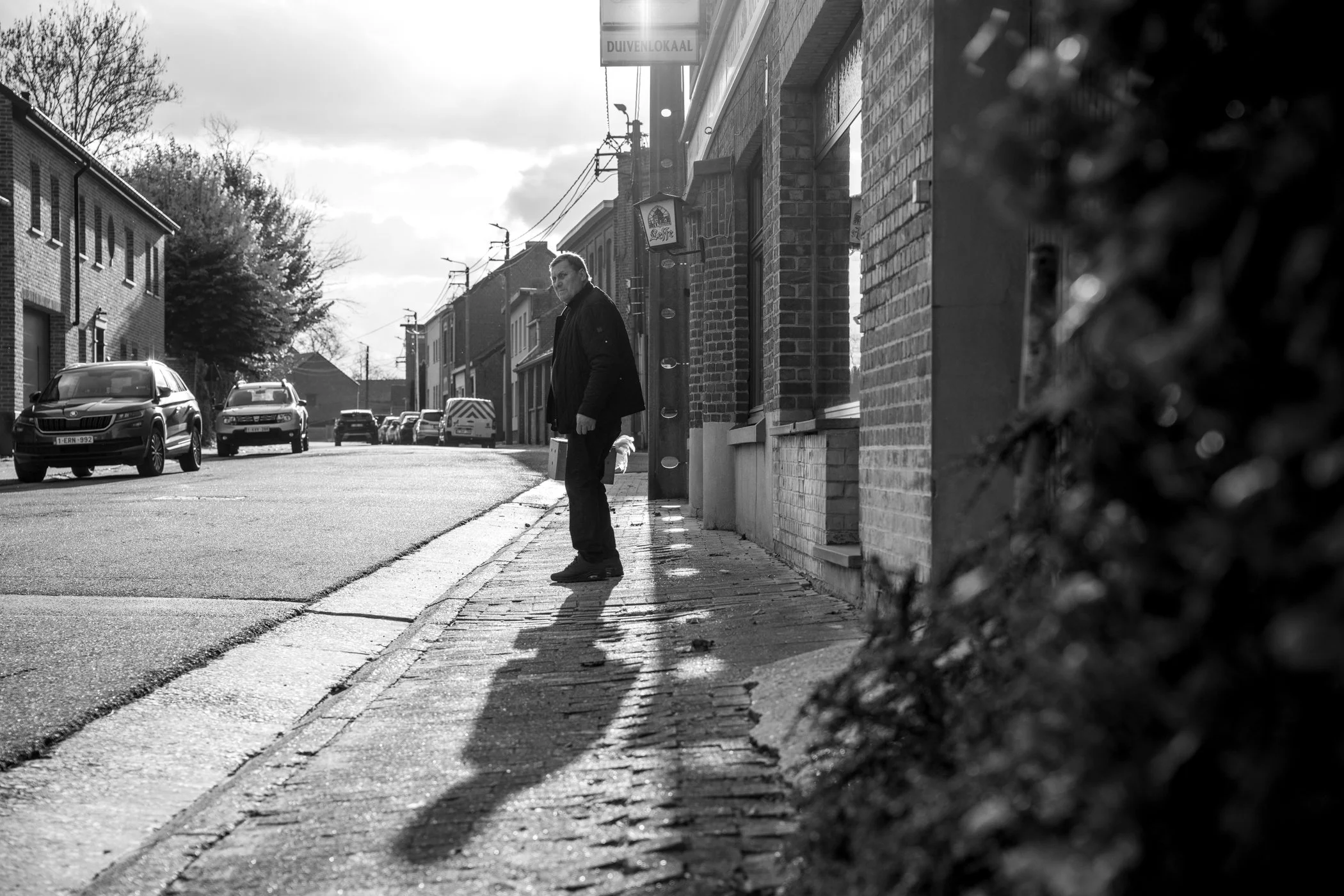 Een man staat op een trottoir met schaduw die op de straat valt, in een rustige straat met geparkeerde auto's en gebouwen.