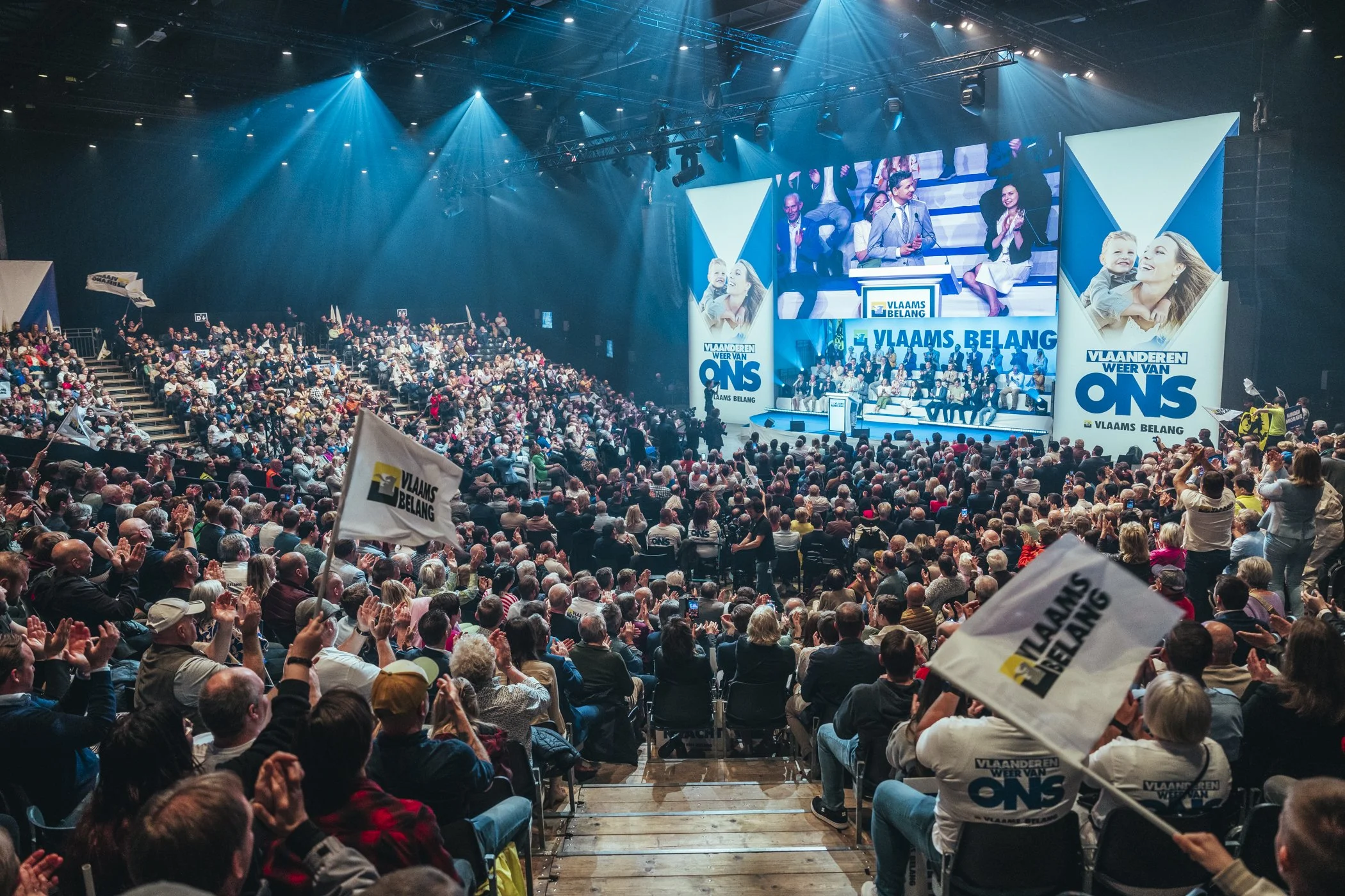 Een grote menigte mensen zit in een auditorium tijdens een evenement. Op het podium staan sprekers en er zijn grote schermen met de tekst 'Vlaams Belang' en 'Vlaanderen weer van ons'. Mensen houden vlaggen en applaudisseren.