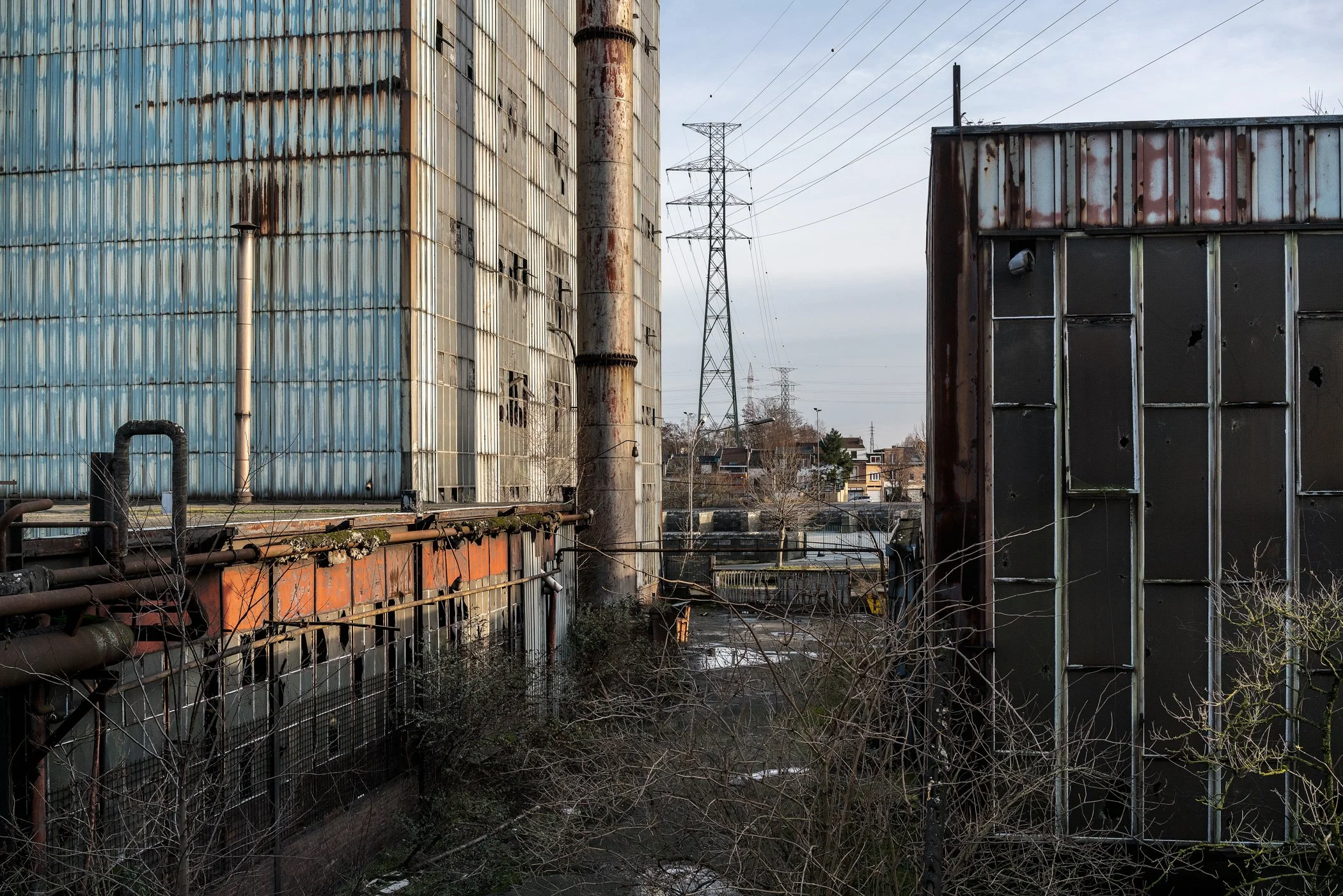 Oud, vervallen fabrieksgebouwen met roestige oppervlakken en doorgebeten kogelgaten in de gevels, met een infrastructuur van elektriciteitsmasten en kabels op de achtergrond.
