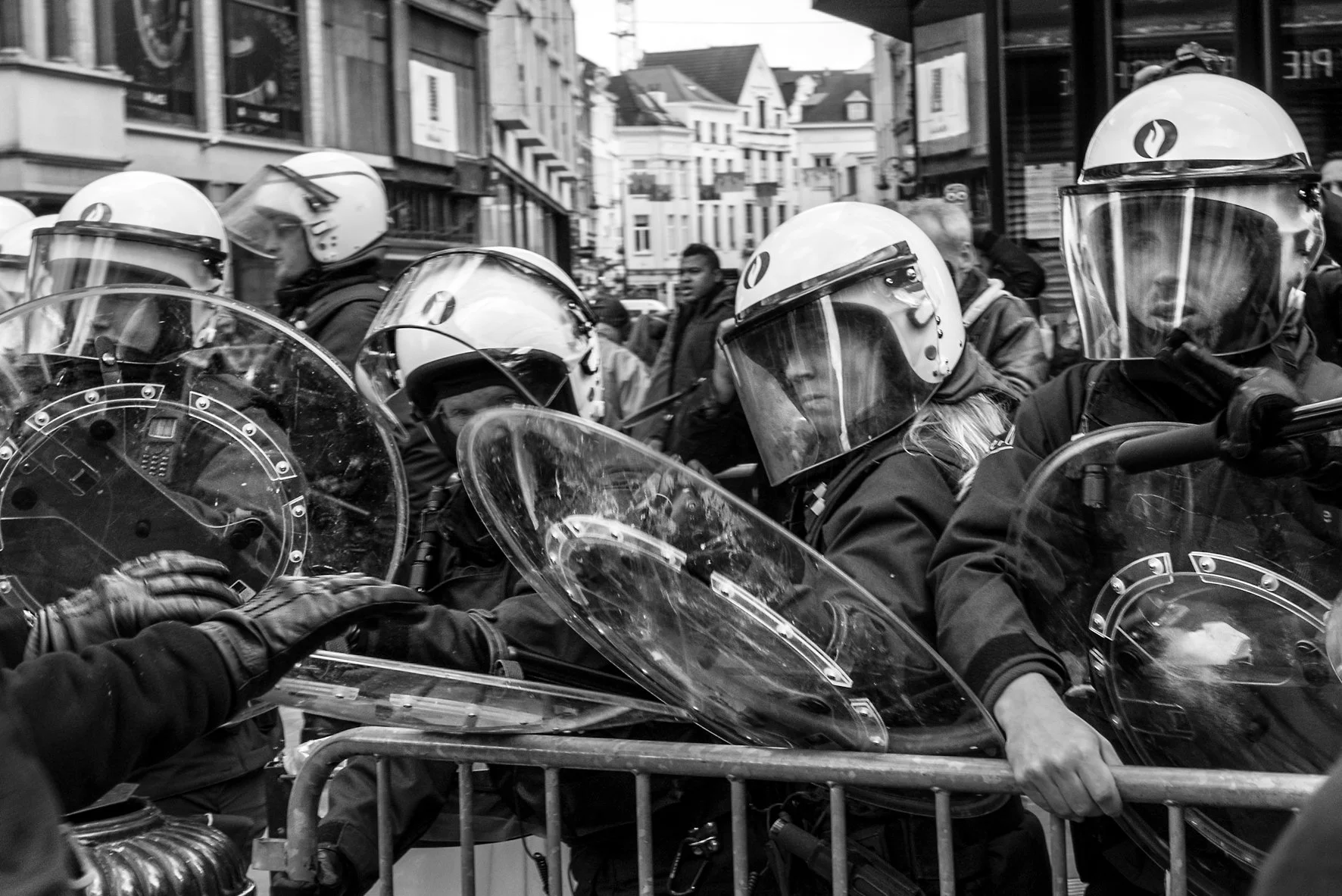 Politie met helm en schild staat in formatie op een stadsstraat, omringd door mensen en gebouwen.