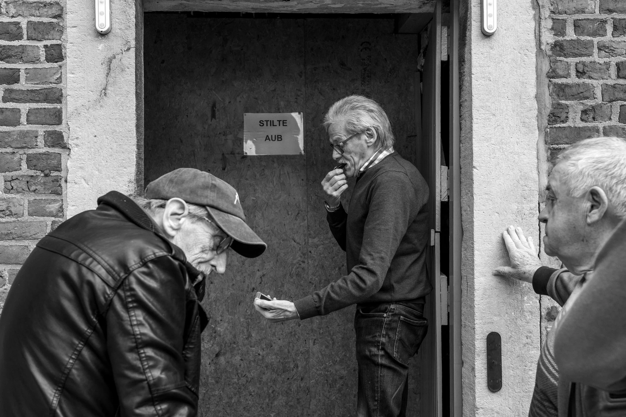 Drie oudere mannen staan buiten een deur, eentje kijkt naar een mobiel apparaat, een kijkt naar een andere man die op de deur klopt en de derde neemt een slok uit een beker.