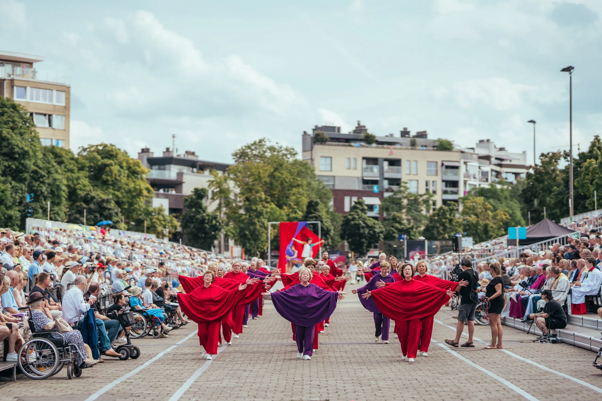Dames in rode en paarse kostuums voeren een dans op een plein uit, omringd door een publiek dat in een openlucht festival zit, met moderne appartementen in de achtergrond.