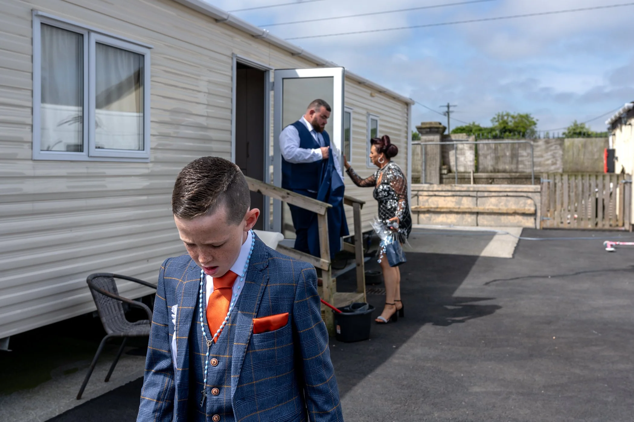 Een jongen in een pak met een oranje stropdas en sieraden kijkt naar beneden. Op de achtergrond staan een man en een vrouw op een trap; de man hoort bij een pak en de vrouw draagt een sexy jurk met pailletten en hakken.