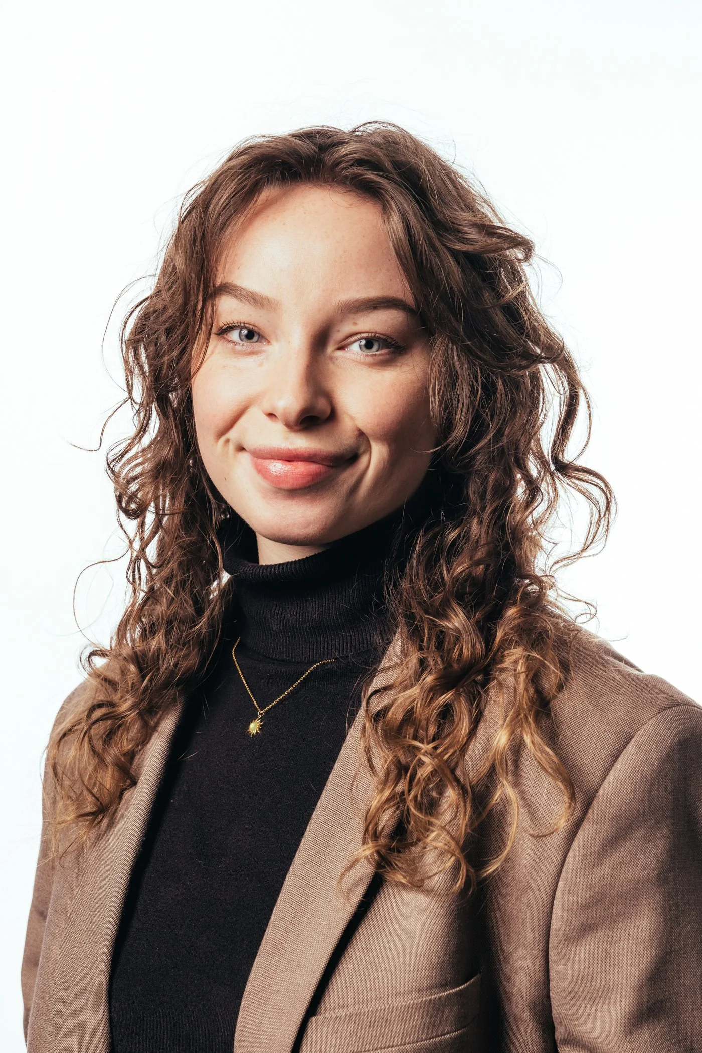 Portret van een jonge vrouw met krullend bruin haar, die glimlacht en gekleed is in een zwarte turtleneck en een beige blazer, met een subtiele gouden ketting met een zonvormige hanger.