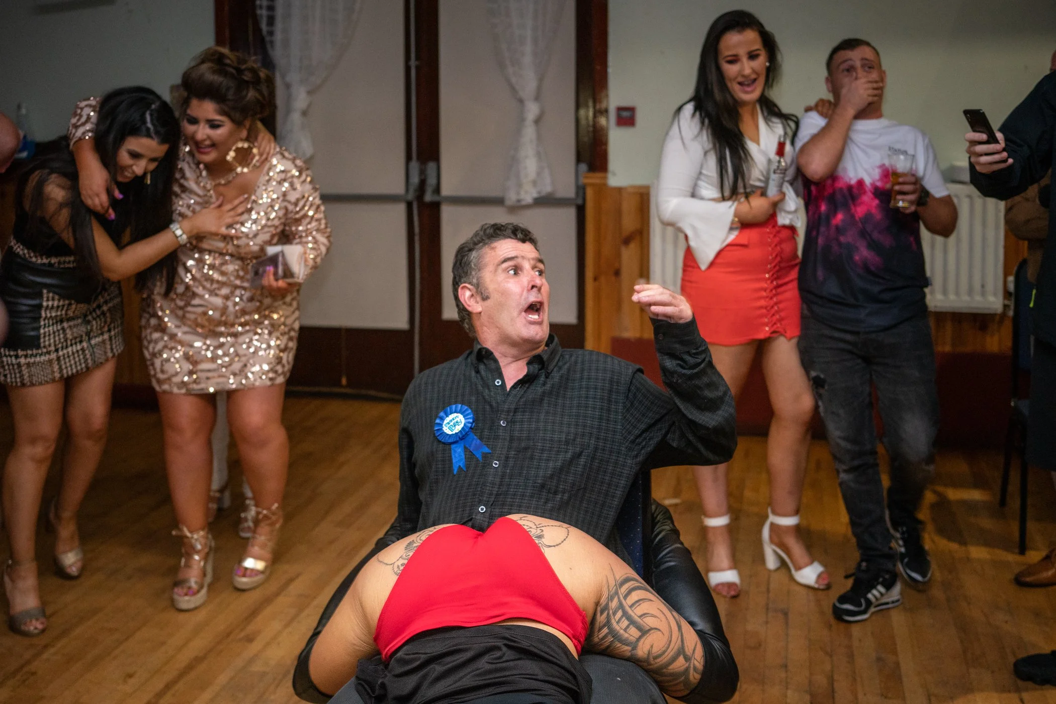 Mensen dansen en lachen op een feestje, man in het midden met een blauwe rozet op zijn overhemd, vrouw in het midden onder de tafel met een rode jurk en tatoeages op haar been, andere mensen in de achtergrond die praten en aan het drinken zijn.