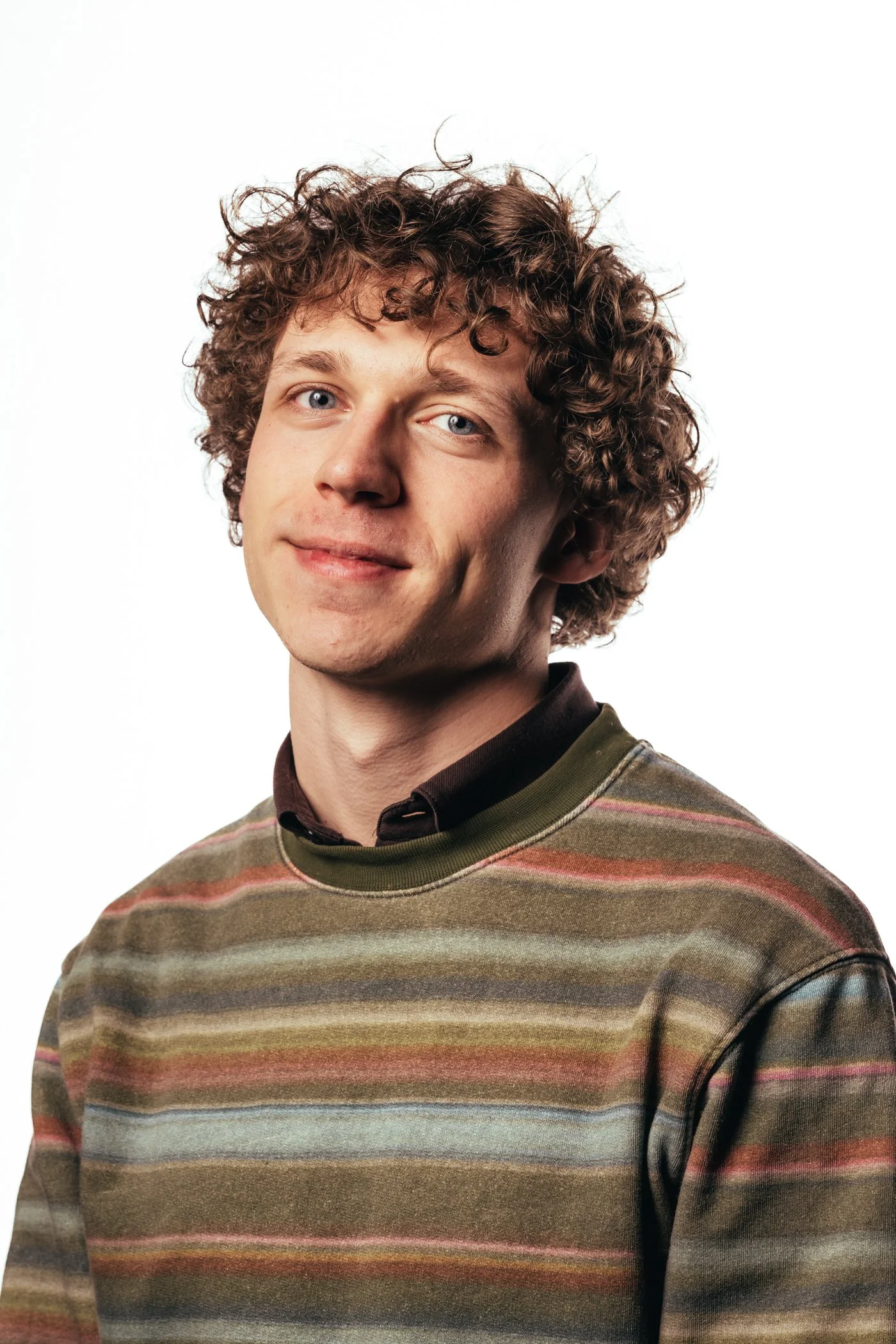 Portret van een jonge man met krullend bruin haar, lichte huid en blauwe ogen, draagt een geruite sweater en een donkere shirt, lachend op een witte achtergrond.