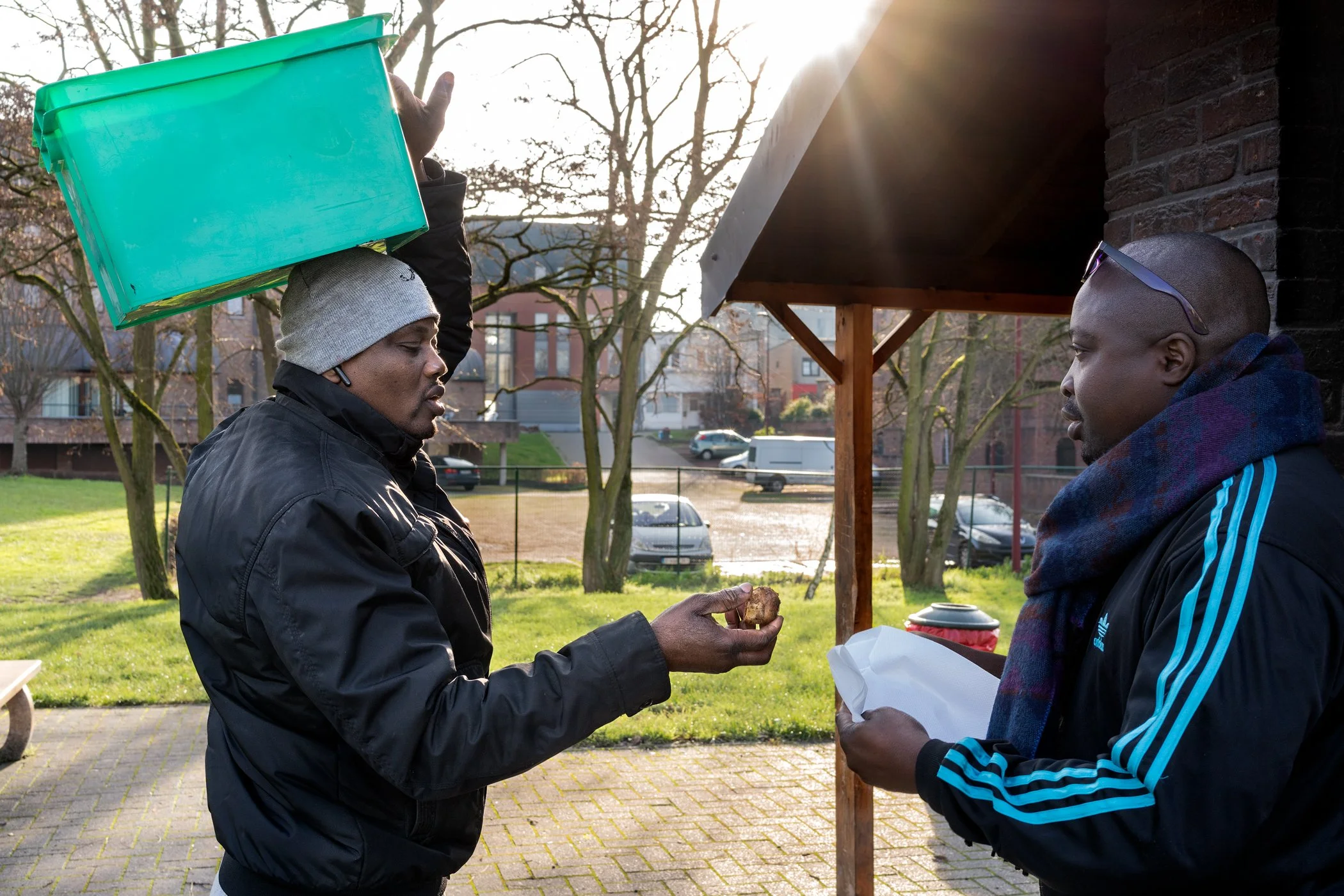 Twee mannen staan buiten bij een marktkastje, een man geeft de ander een stuk voedsel terwijl de zon ondergaat.