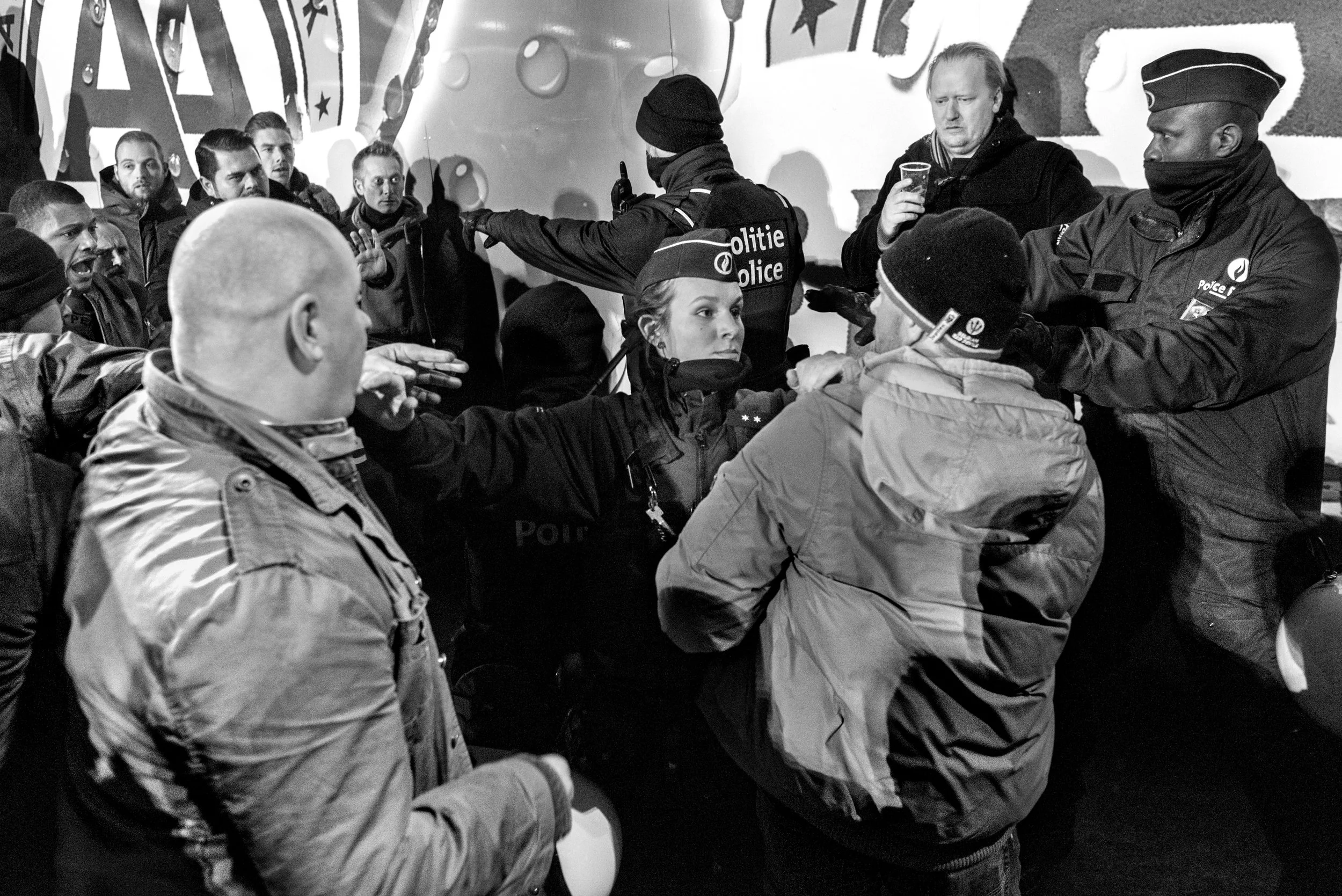 Politieagenten en burgers in een confrontatie, met politieuniformen en een muur met opvallende graffiti op de achtergrond.