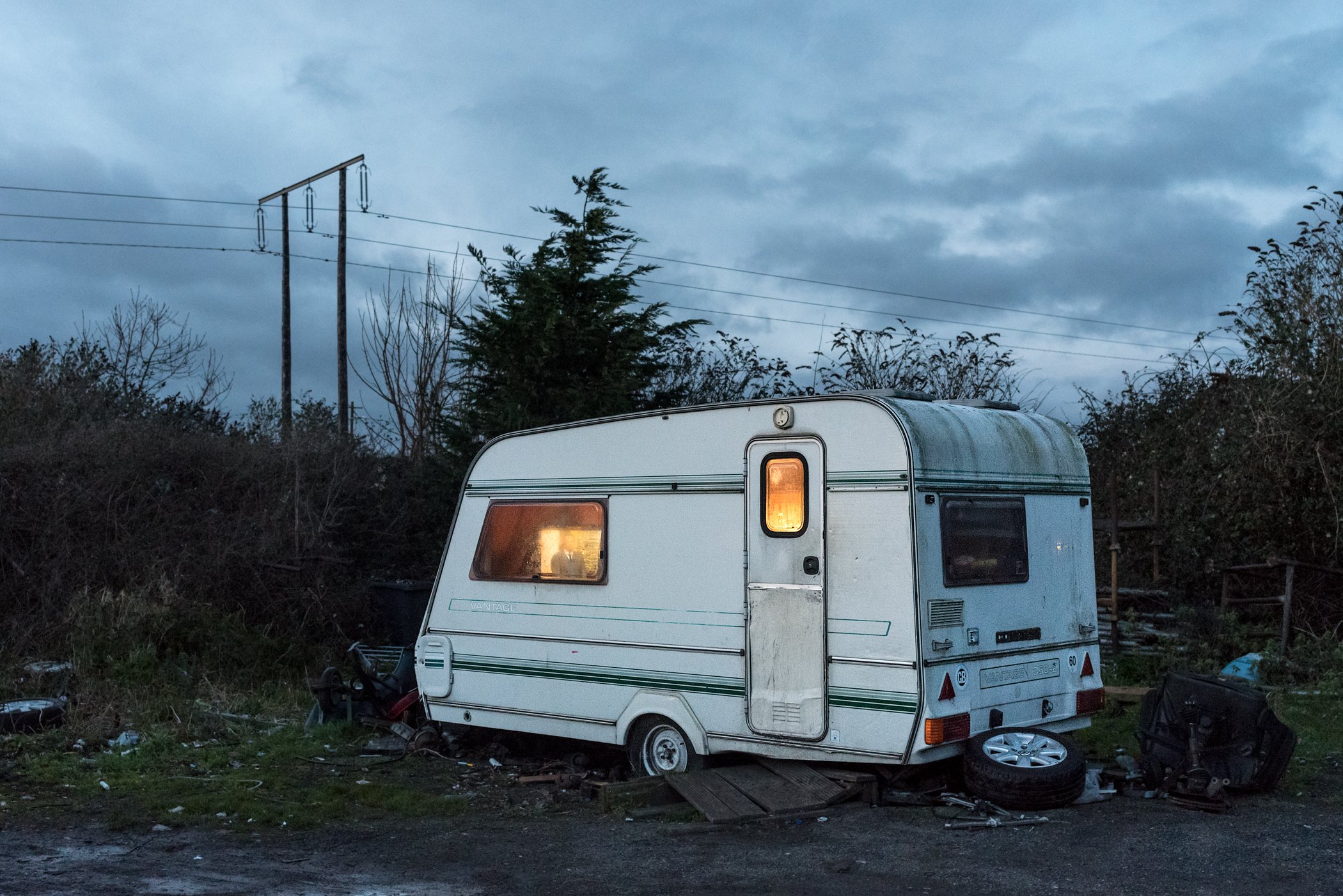 Een oude, verwarde caravan op een vuilige plek, omringd door afval, tegen een bewolkte hemel met hoogspanningsdraden op de achtergrond.