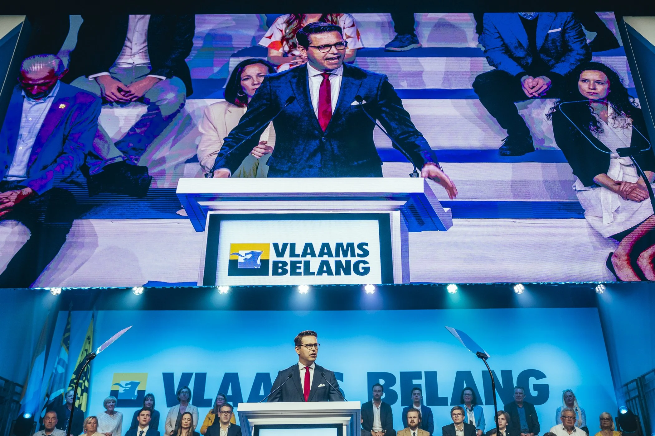 Manen en vrouwen op een podium bij een politieke bijeenkomst met het logo 'Vlaams Belang' op de achtergrond, een man in een zwart pak spreekt achter een witte lessenaar.
