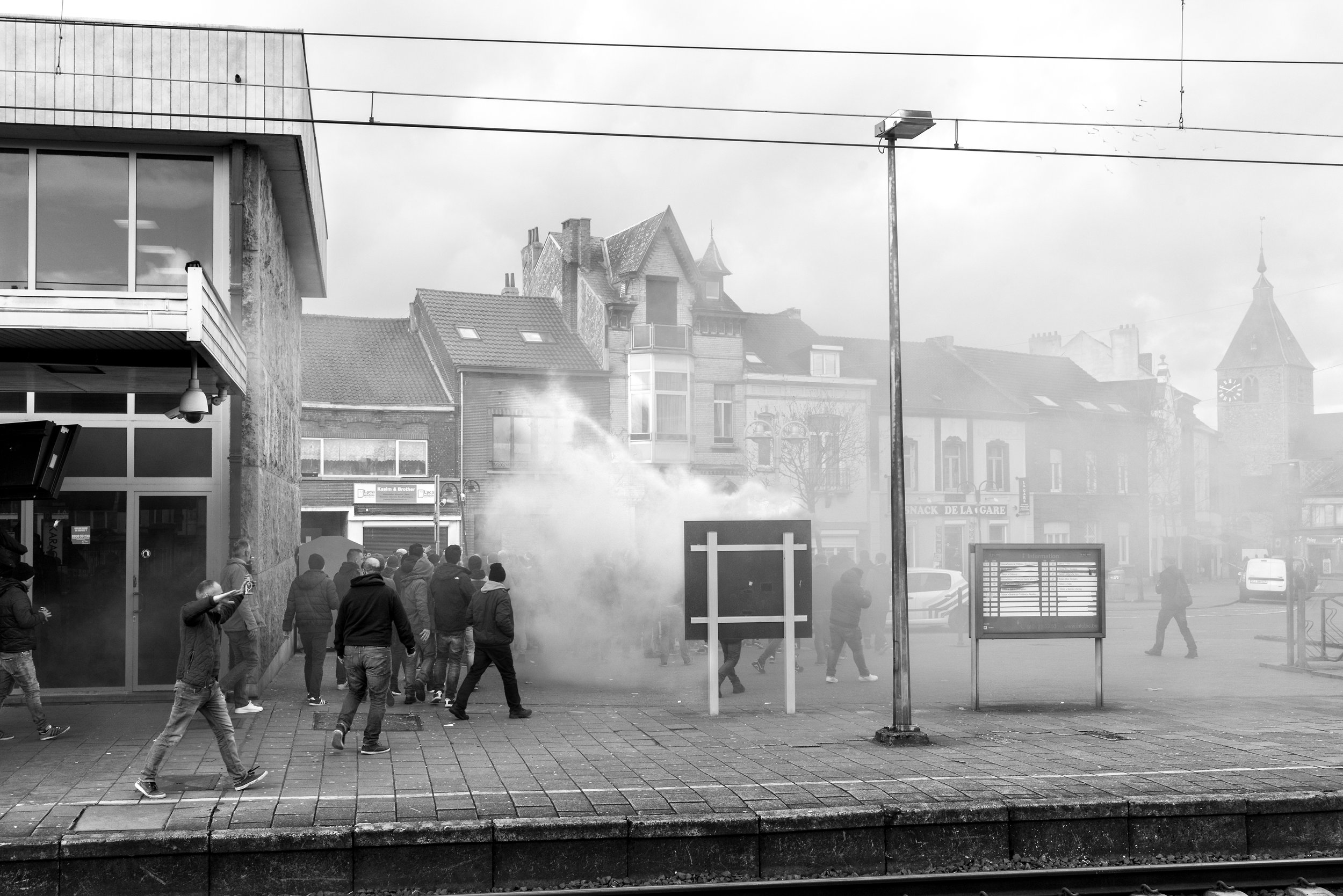 Een groep mensen op een plein met rook of mist. Er is een bushokje, lampen, en gebouwen op de achtergrond, waaronder een kerktoren en een winkeltje.