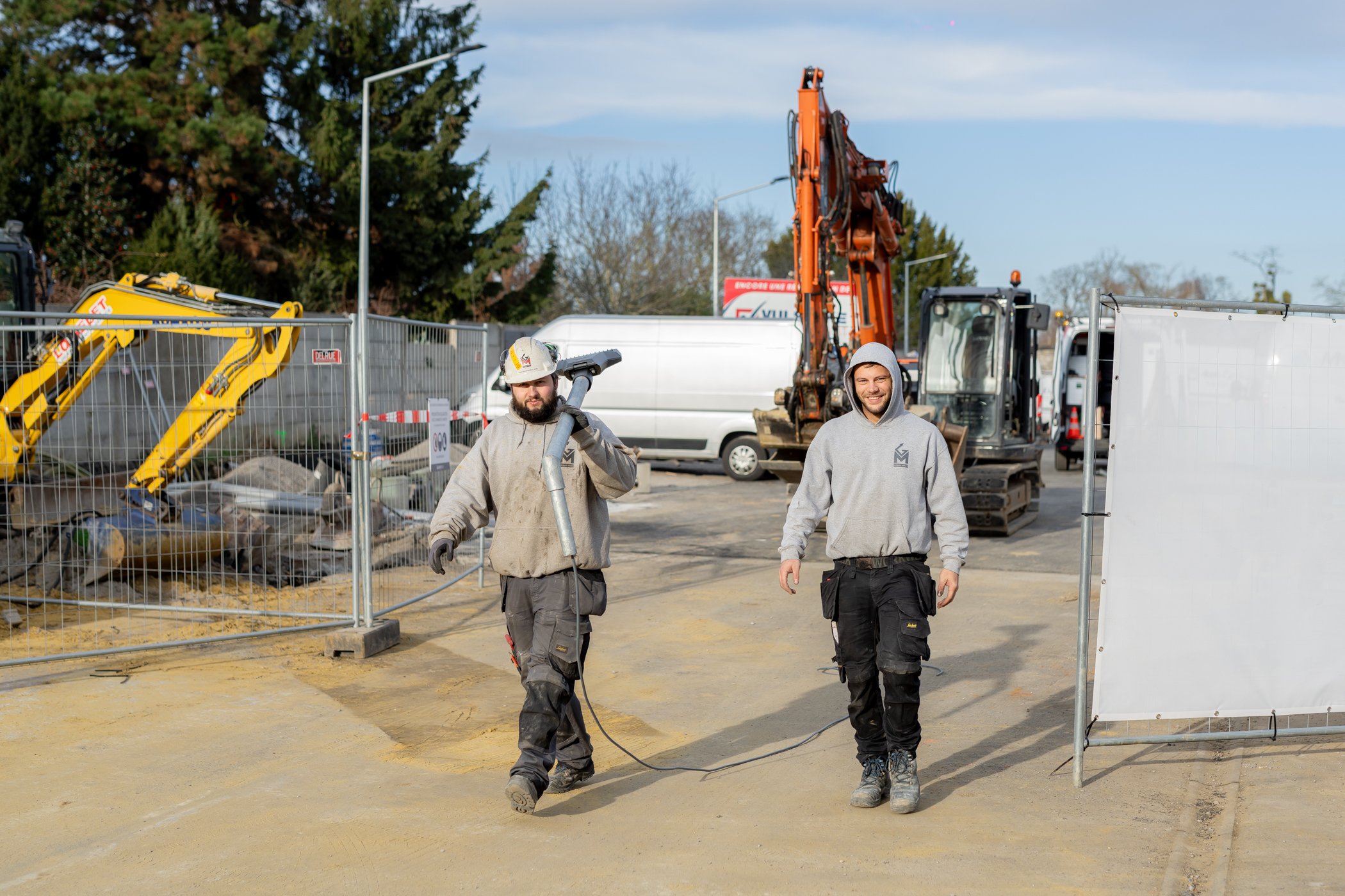 Twee bouwvakkers lopen op een bouwplaats, een van hen draagt een veiligheidshelm en houdt een elektrische boor, de andere lacht en draagt een cap. In de achtergrond staan voertuigen en bouwmachines.